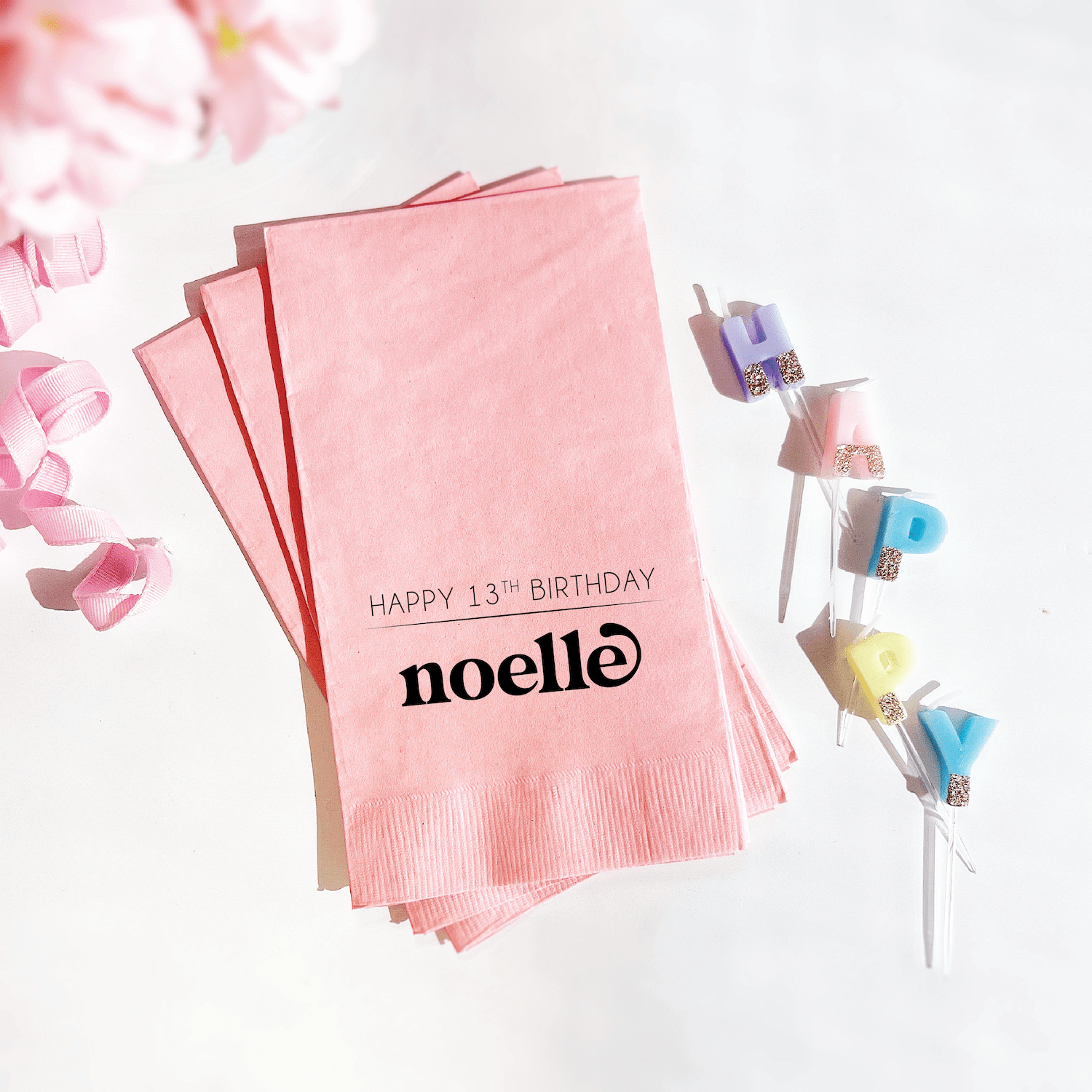 Stack of pink napkins with "Happy 13th Birthday" and "noelle" foil stamped text, displayed with colorful lollipops and flower petals.