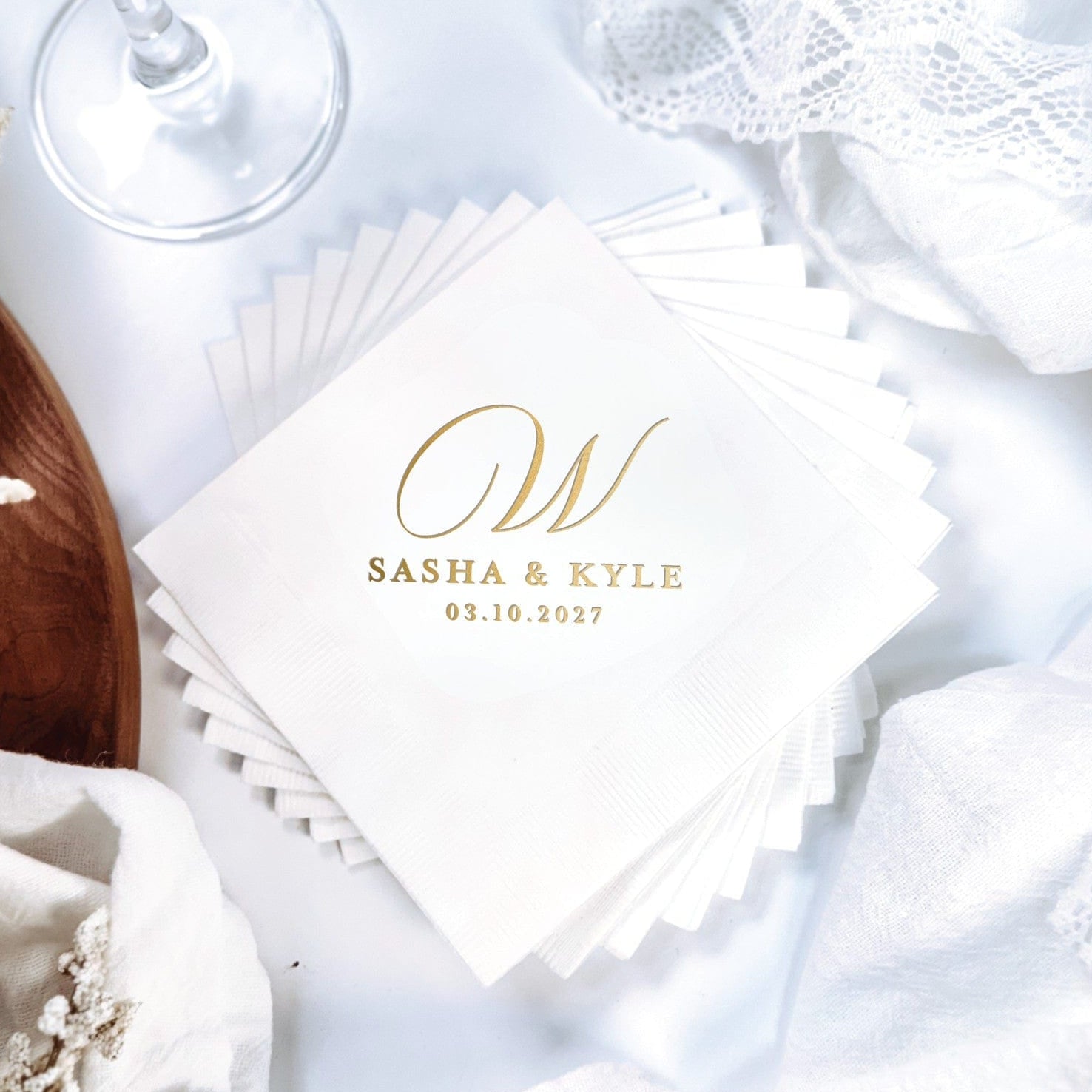 A stack of elegant white disposable wedding napkins featuring a custom metallic gold foil initial design, set amidst delicate white fabric and floral accents for a special event.