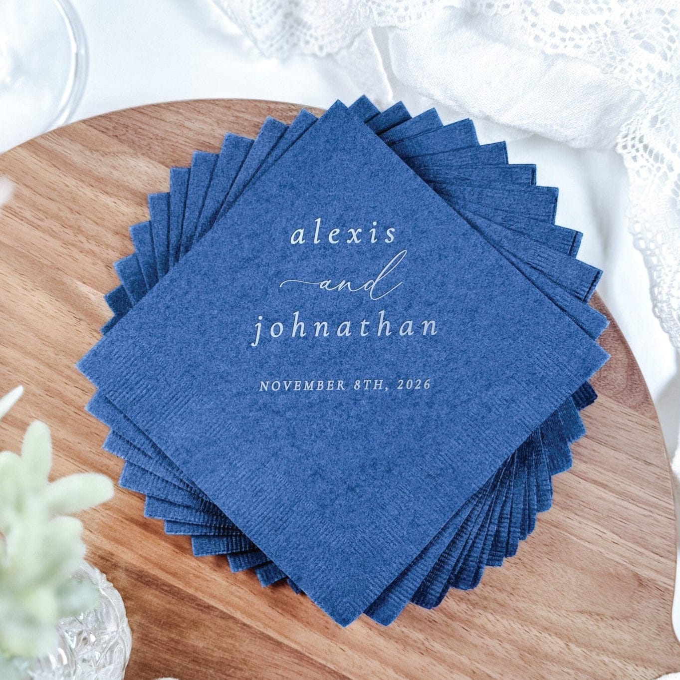 A stack of personalized navy blue disposable paper napkins with white print, arranged on a wooden serving board alongside a delicate white lace fabric and a champagne glass, ideal for a wedding or elegant event.
