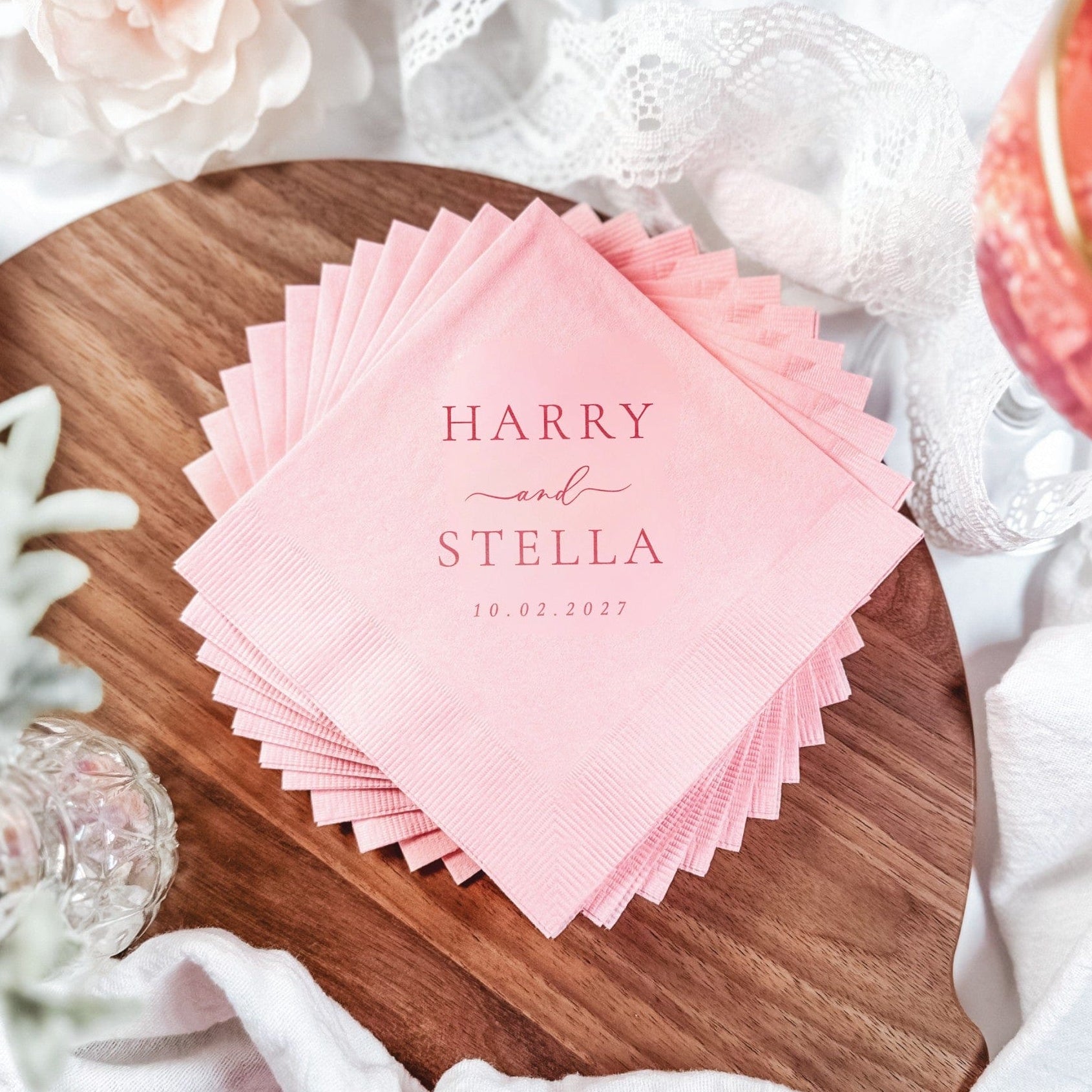 A stack of pale pink paper napkins with a subtle textured edge, personalized with a custom design, arranged on a wooden board for a wedding celebration.