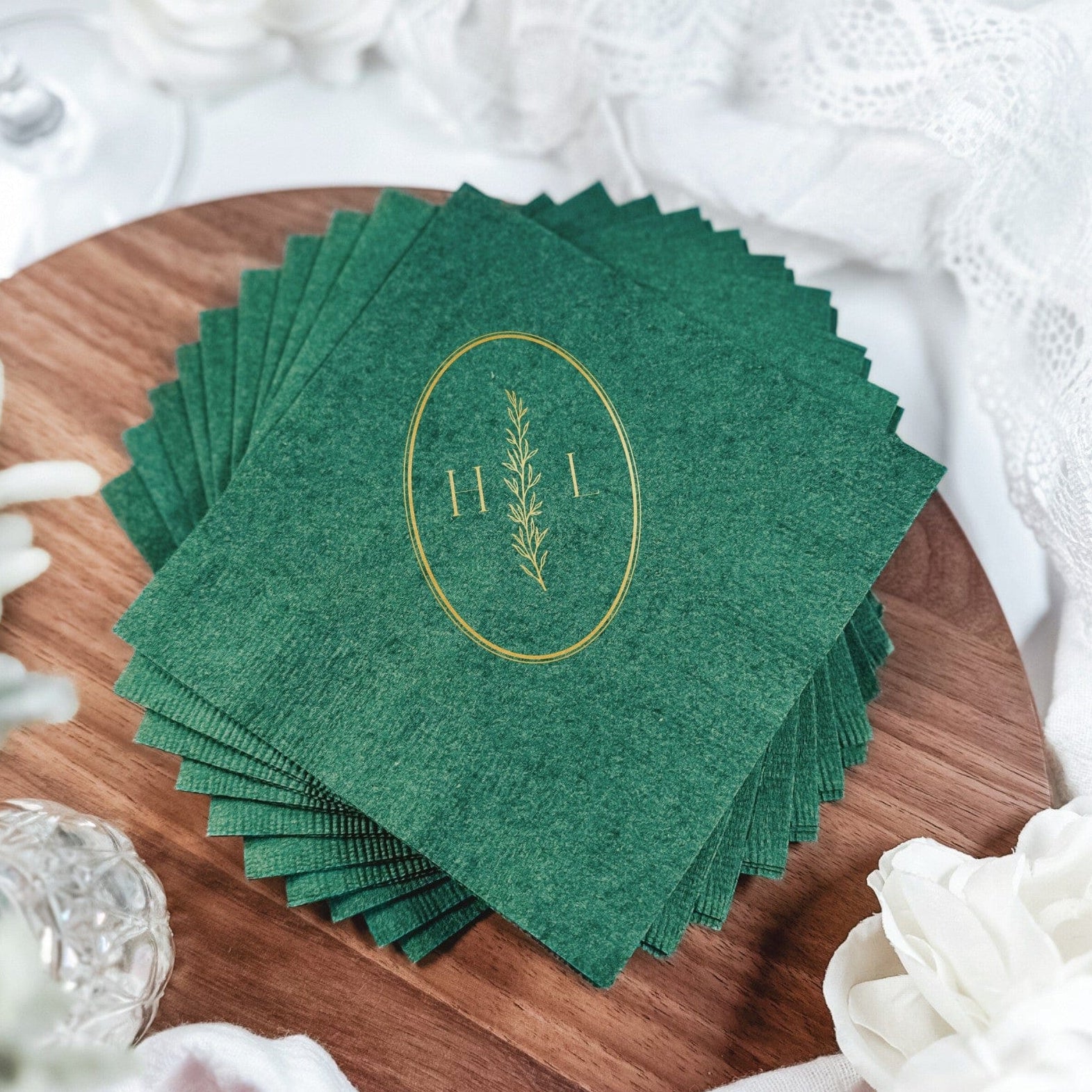A stack of deep green paper napkins featuring a gold foil oval design with a botanical branch, perfect for wedding or elegant event table settings.