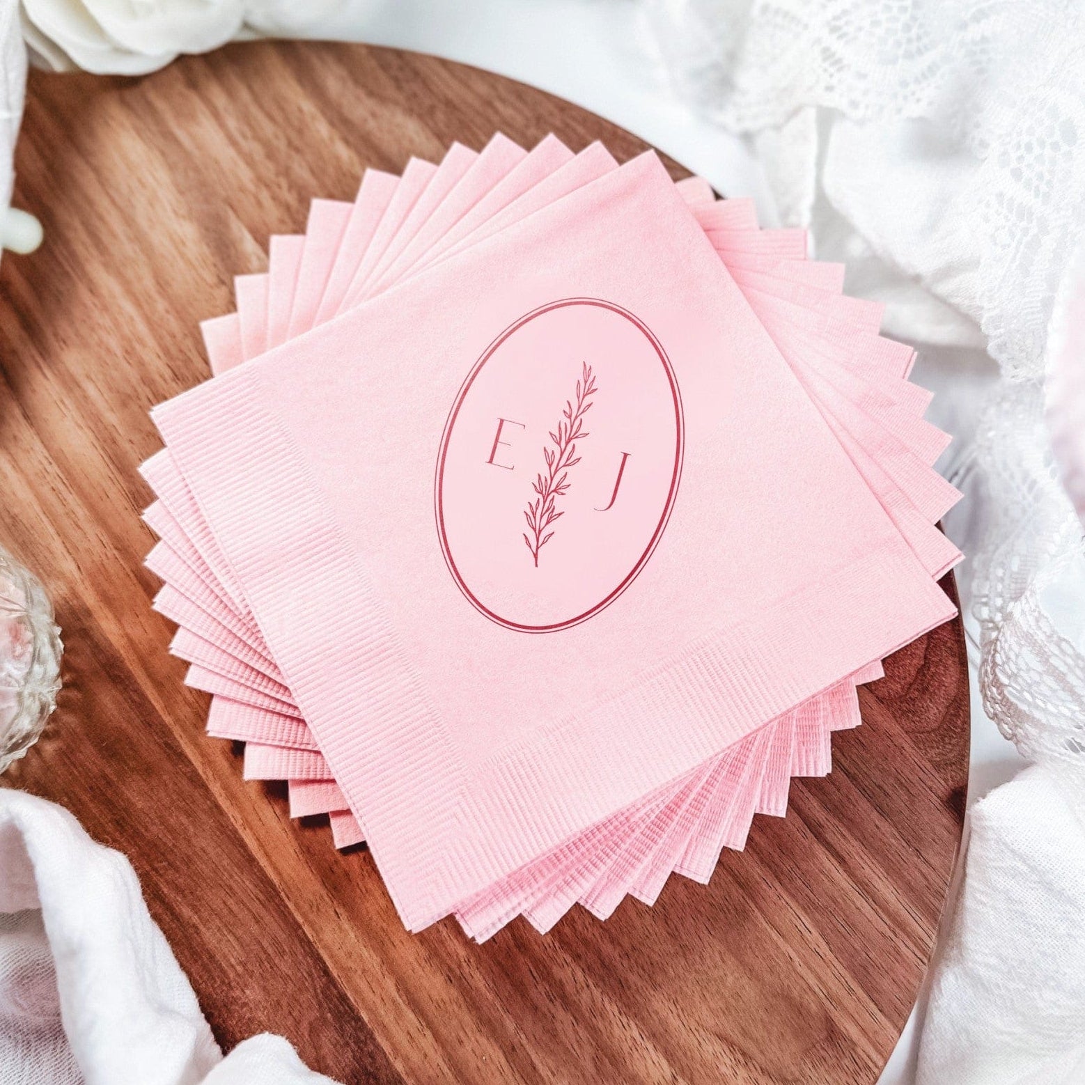 A stack of light pink paper napkins is fanned out on a wooden board, each personalized with a darker pink oval design featuring a botanical sprig and two initials, ideal for wedding or event decor.