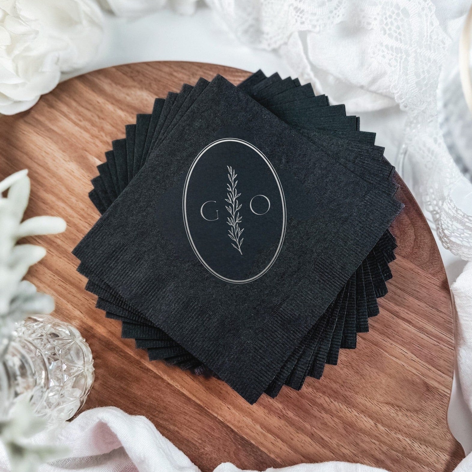 Elegant black paper wedding napkins personalized with a silver foil design featuring an oval frame and a delicate botanical branch, perfect for event tablescapes.