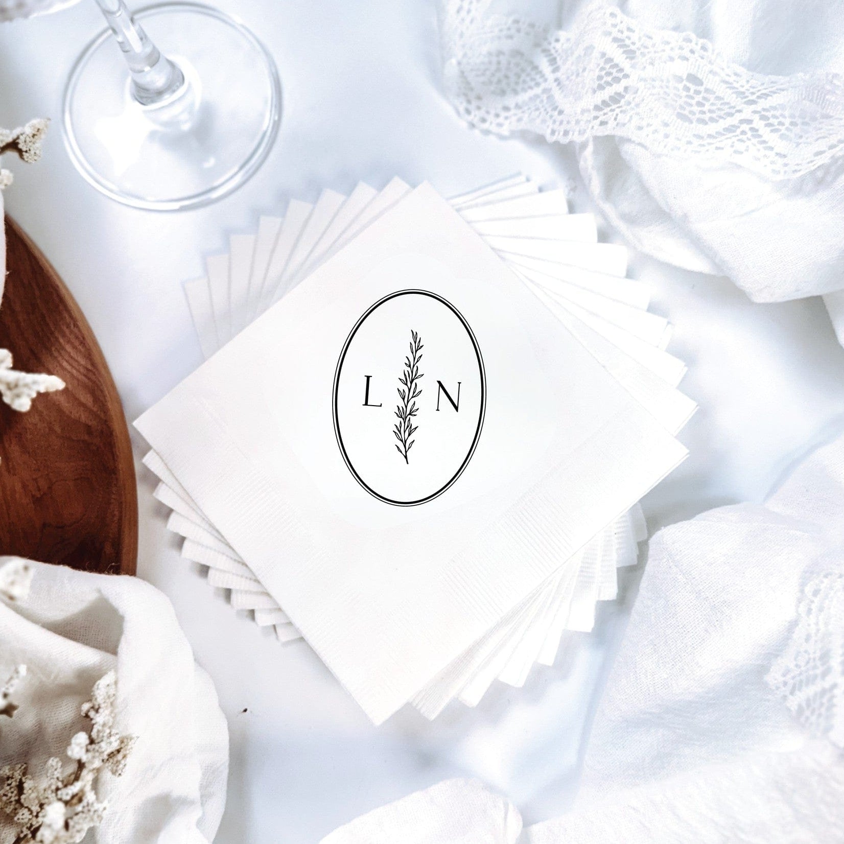 A stack of white paper wedding napkins is personalized with an elegant black oval design featuring a botanical sprig, perfect for a refined event.