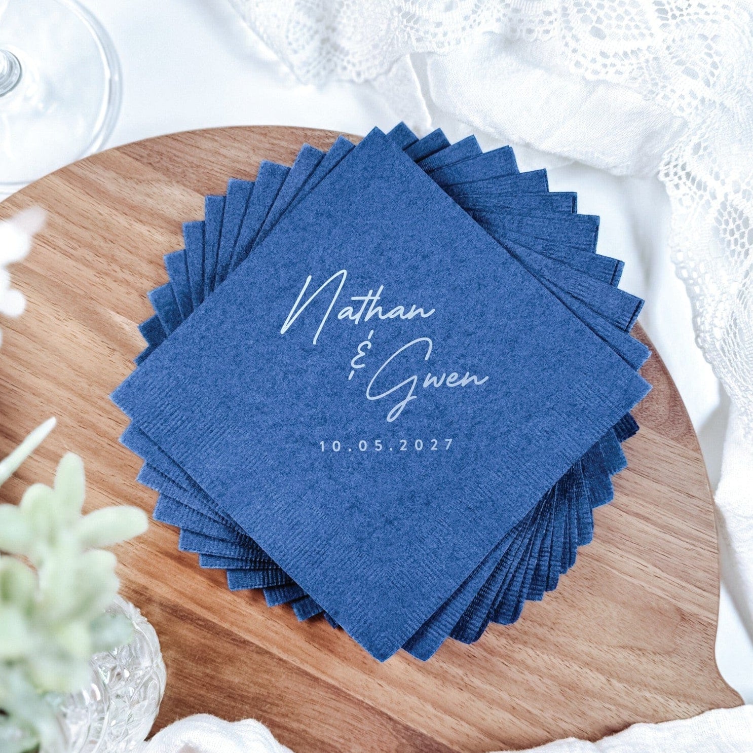A stack of navy blue paper wedding napkins featuring personalized white text, neatly arranged on a wooden board amidst white lace and greenery.