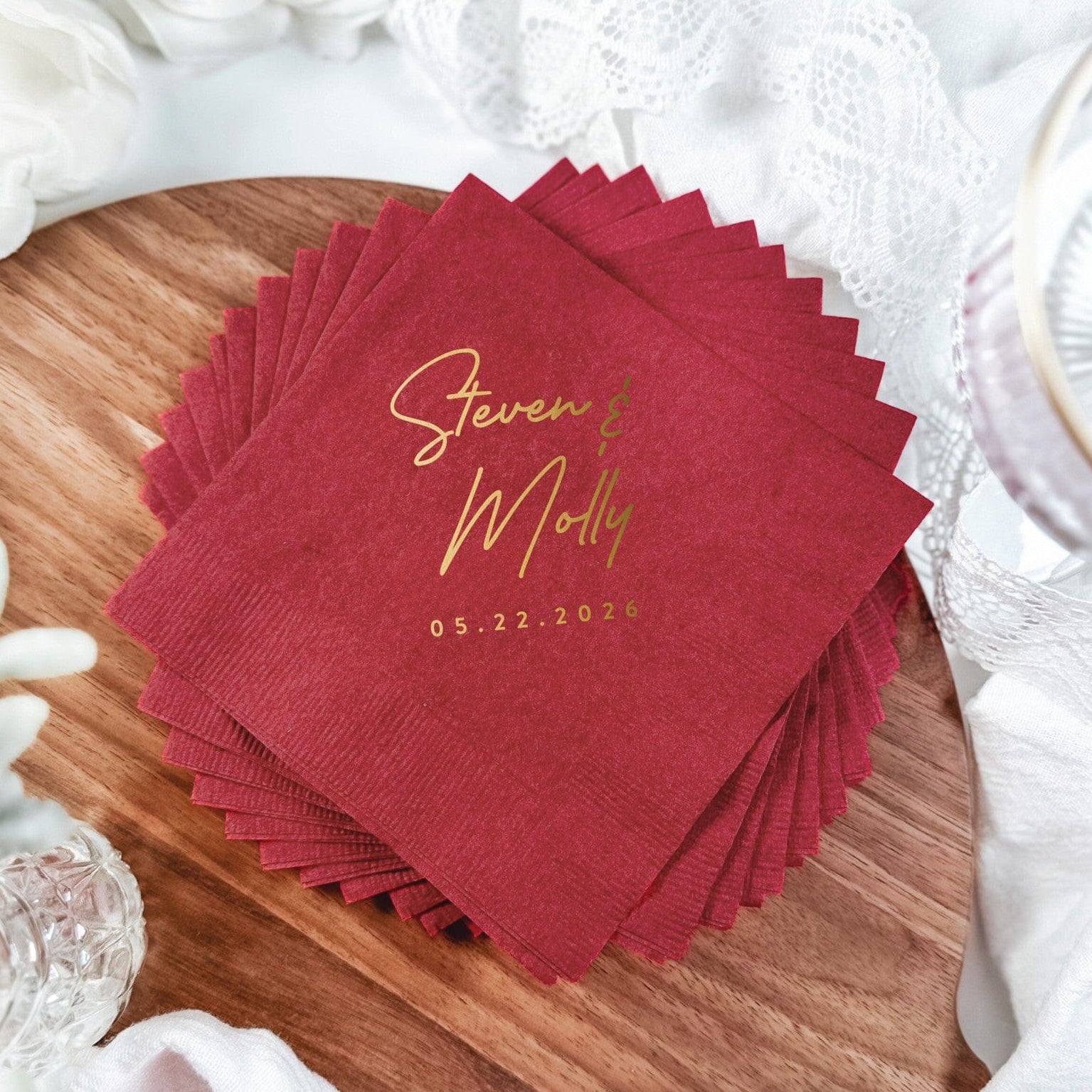 A stack of deep red disposable paper napkins featuring elegant gold foil script, presented on a wooden board with white lace and flowers, ideal for a wedding or sophisticated event.