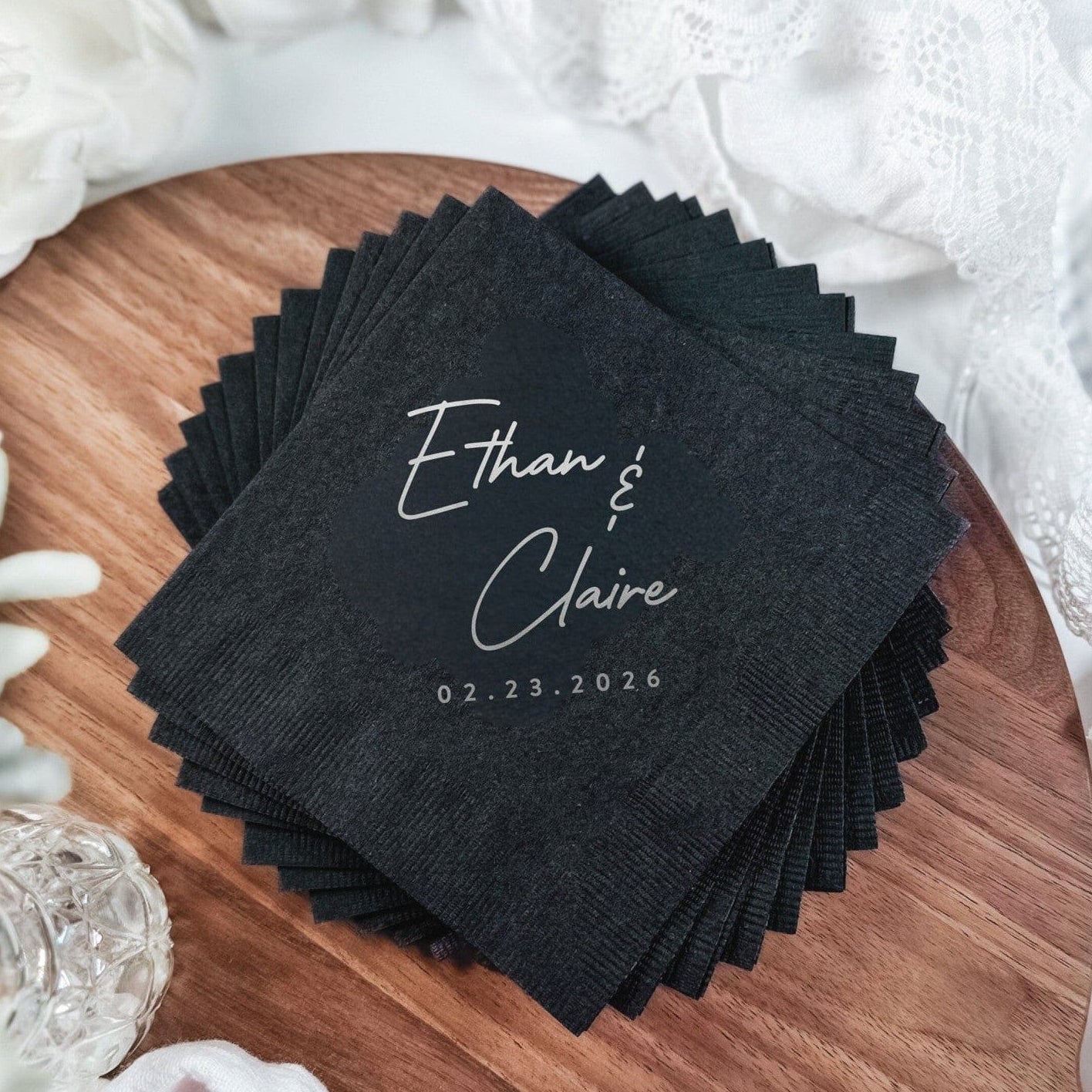 A stack of black textured paper beverage napkins personalized with elegant white script font, displayed on a wooden board with white lace and flowers, ideal for a wedding.