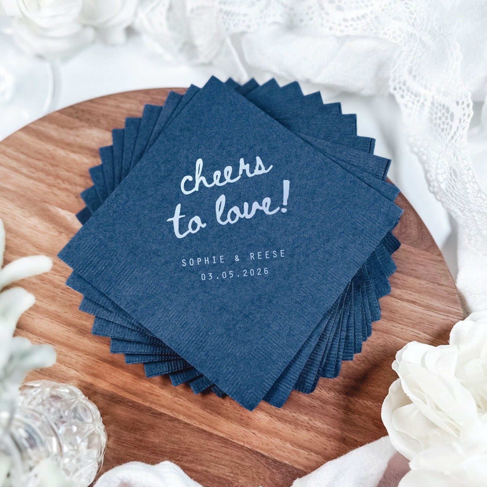 A stack of navy blue square paper napkins with scalloped edges is displayed on a rustic wooden board, suitable for a wedding or event.