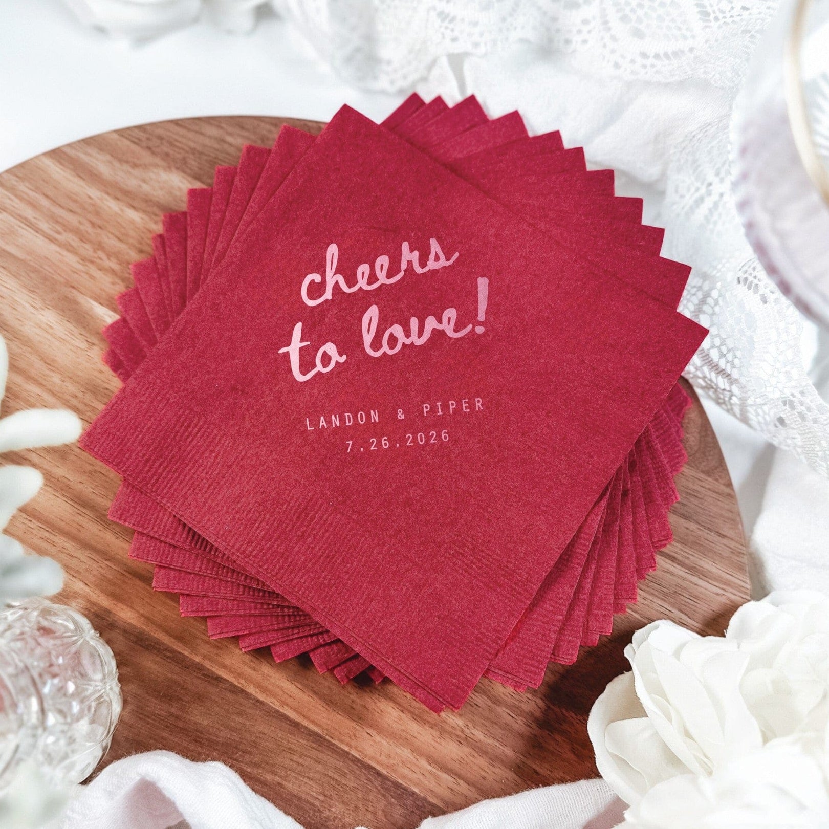 A stack of deep red square paper napkins with a subtle textured finish and scalloped edges, featuring a pale pink 'cheers to love!' design, ideal for a wedding or celebratory event.