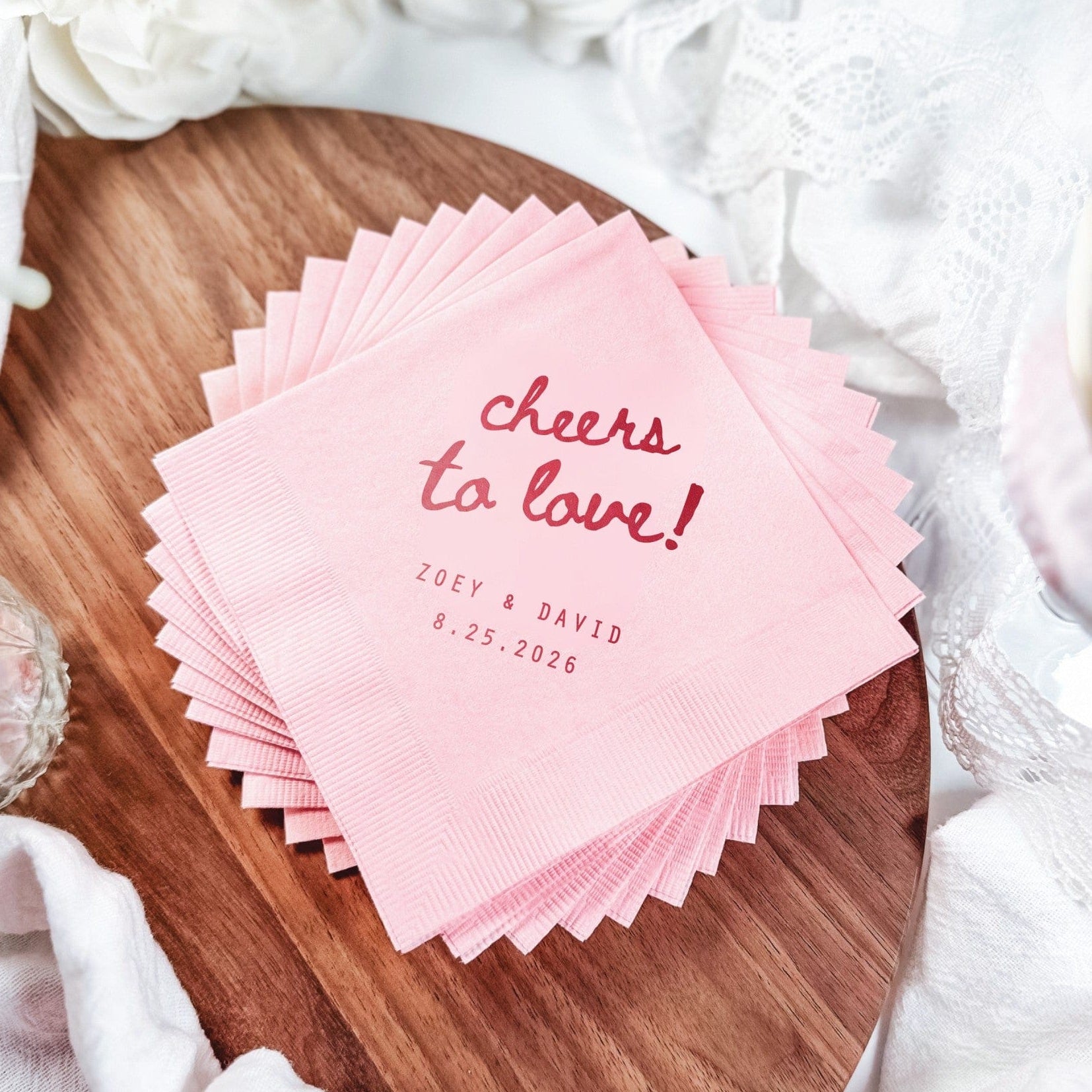A stack of light pink personalized paper wedding napkins featuring a festive 'cheers to love!' design, perfect for a celebratory event.