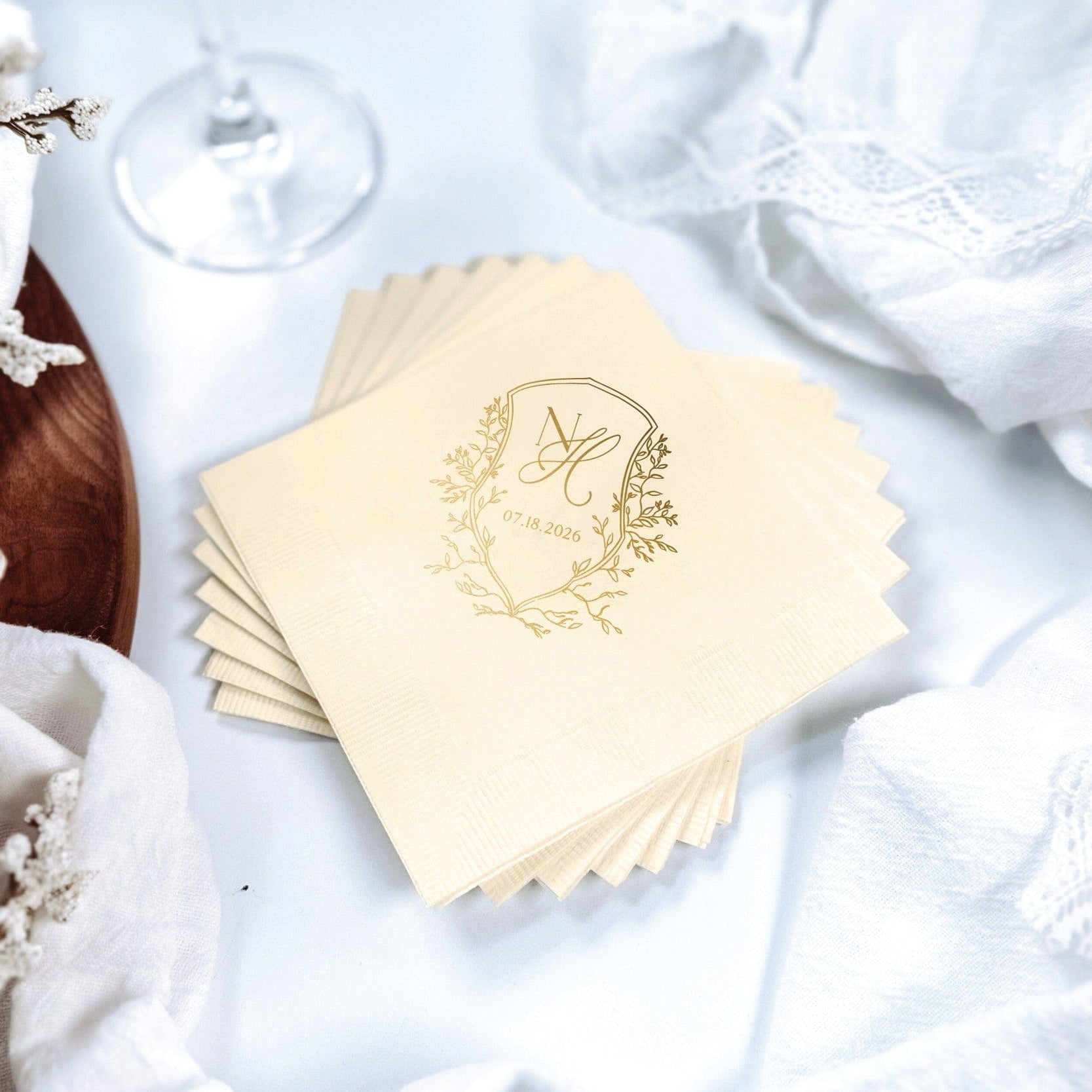 A stack of light cream wedding napkins featuring a gold foil printed crest design with a monogram and date placeholder, perfect for personalized event table decor.