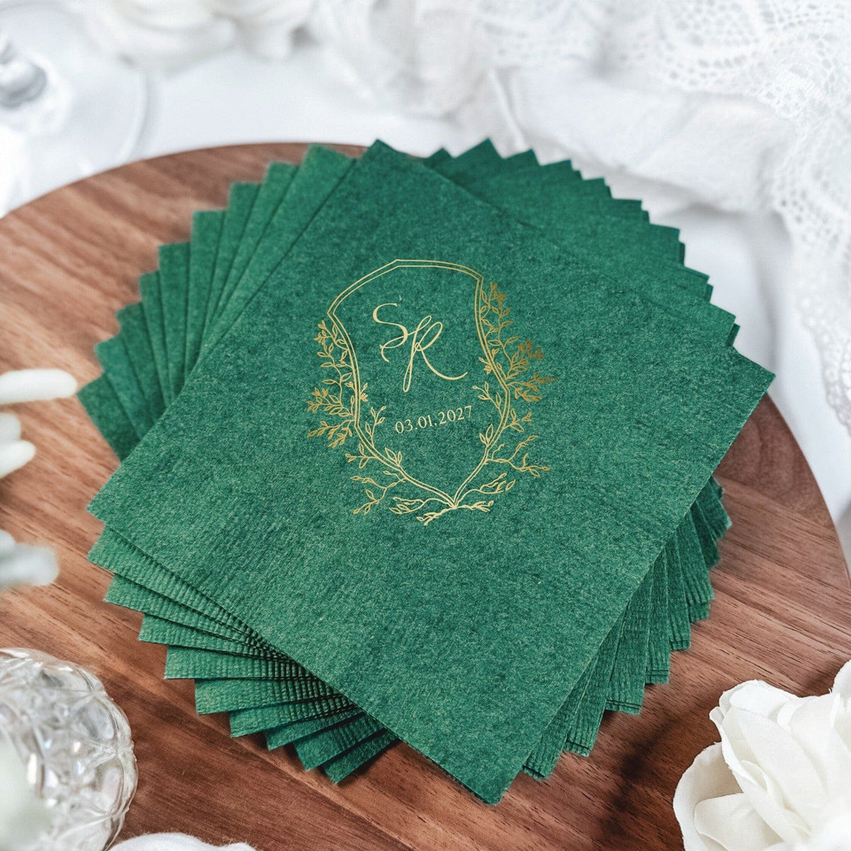 A stack of deep forest green paper napkins featuring a personalized gold foil monogram set within a foliage wreath design, ideal for a wedding or elegant event.