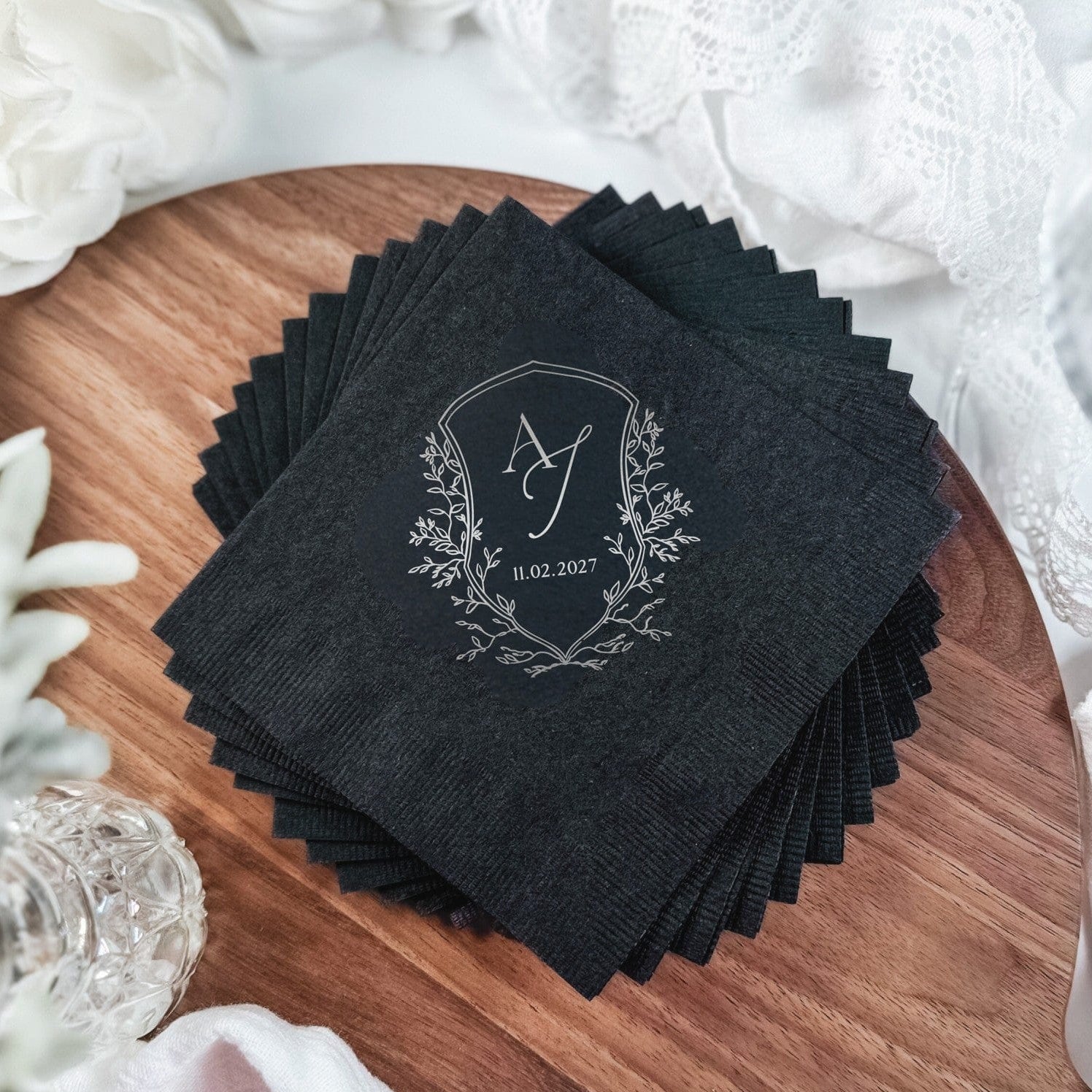 A stack of dark gray paper wedding napkins featuring a silver foil personalized crest design with a monogram, perfect for event decor.