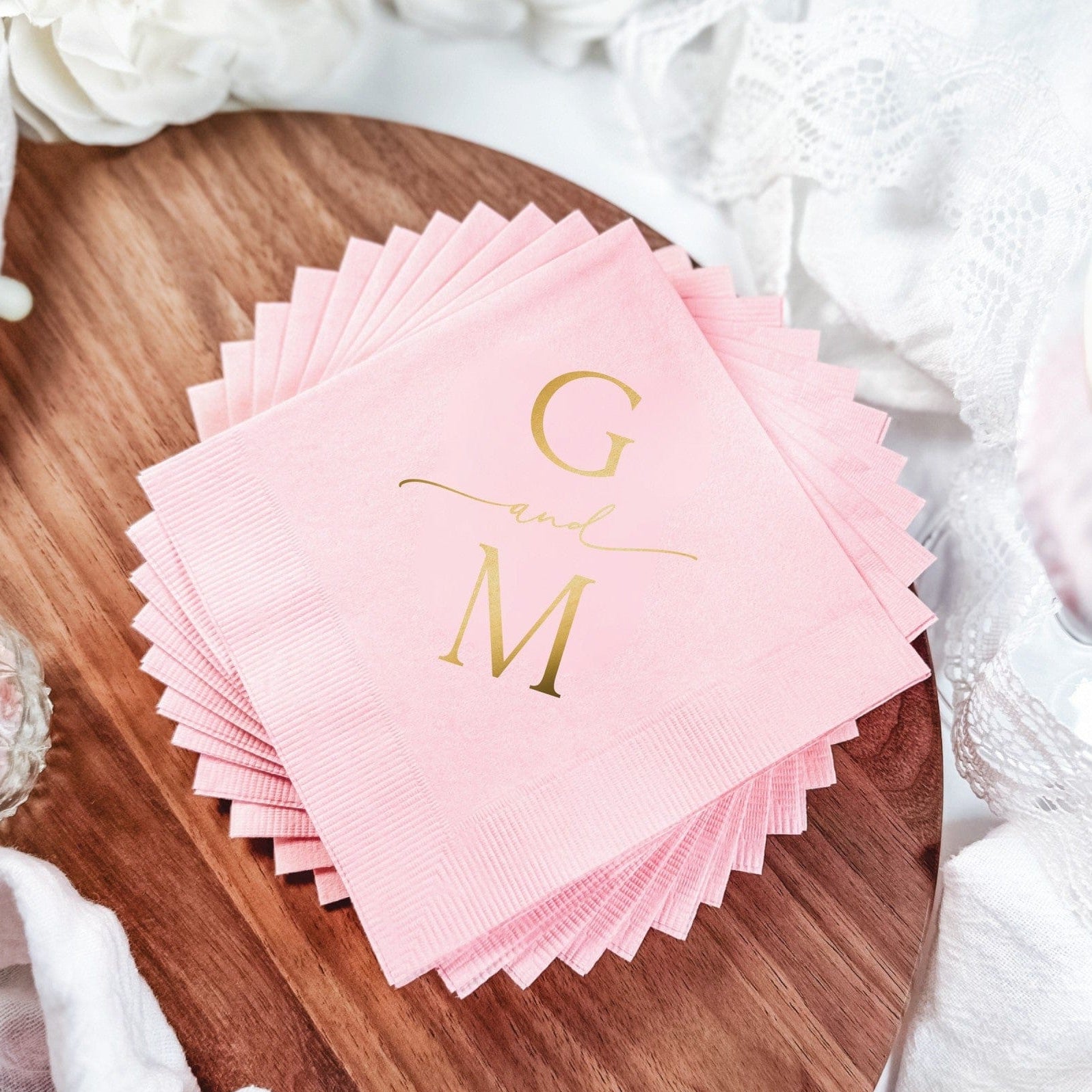 A stack of textured light pink disposable paper napkins featuring elegant gold foil monogramming, suitable for a wedding or sophisticated event setting.