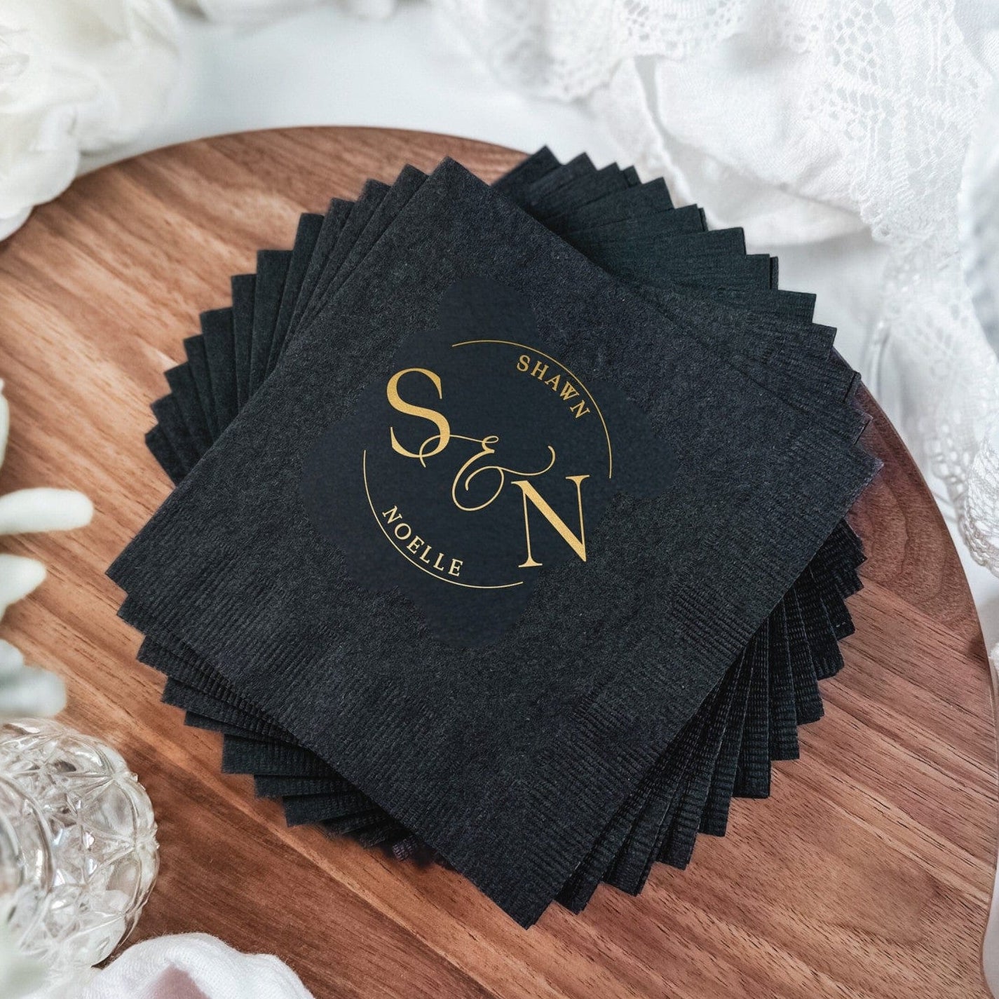 A stack of black disposable wedding napkins featuring a custom metallic gold monogram design, elegantly displayed on a wooden board with white lace and floral accents.