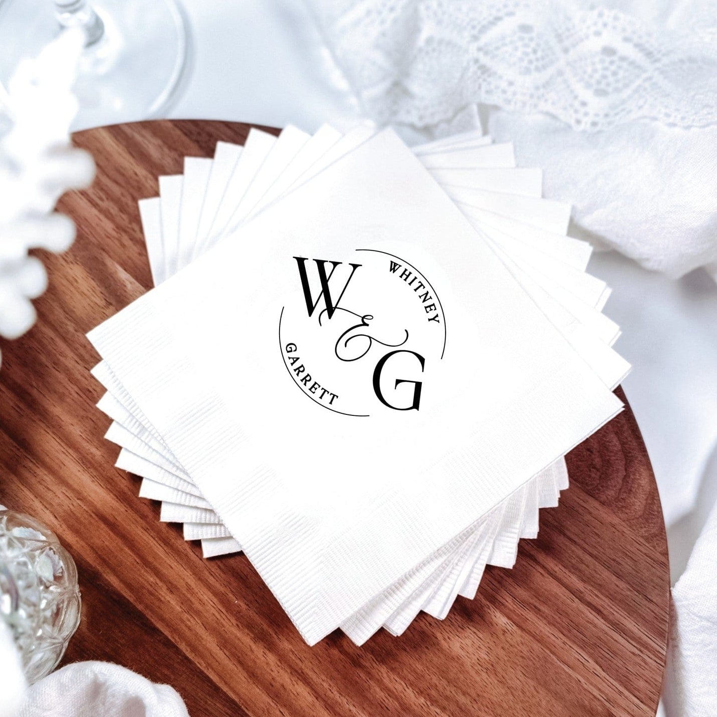 A stack of elegant white textured paper wedding napkins, personalized with a custom initial monogram design, artfully arranged on a rustic wooden board.