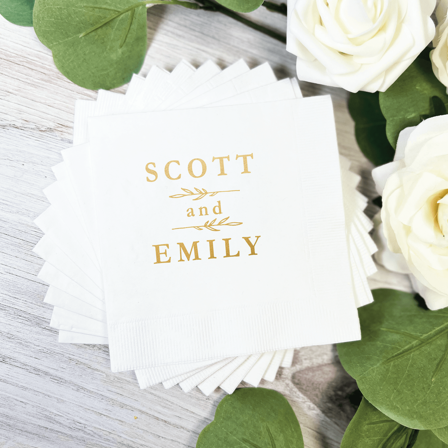 White cocktail napkins with gold foil-stamped names "Scott and Emily" with botanical leaf design, surrounded by white roses and greenery.