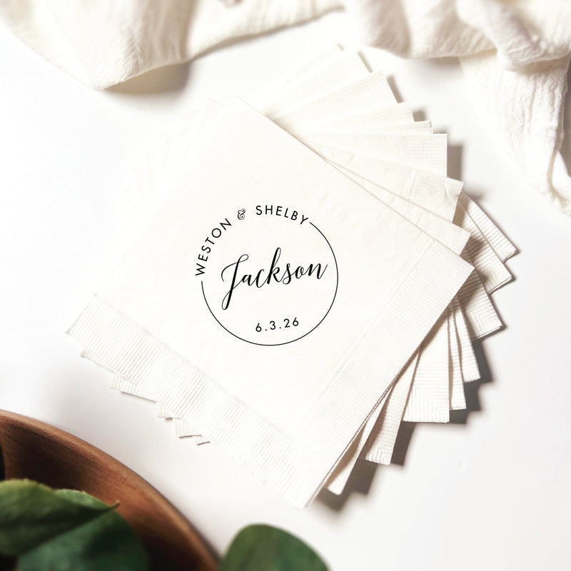 A stack of white disposable napkins, made of soft paper, featuring a custom printed design within a circular border for a wedding or special event.
