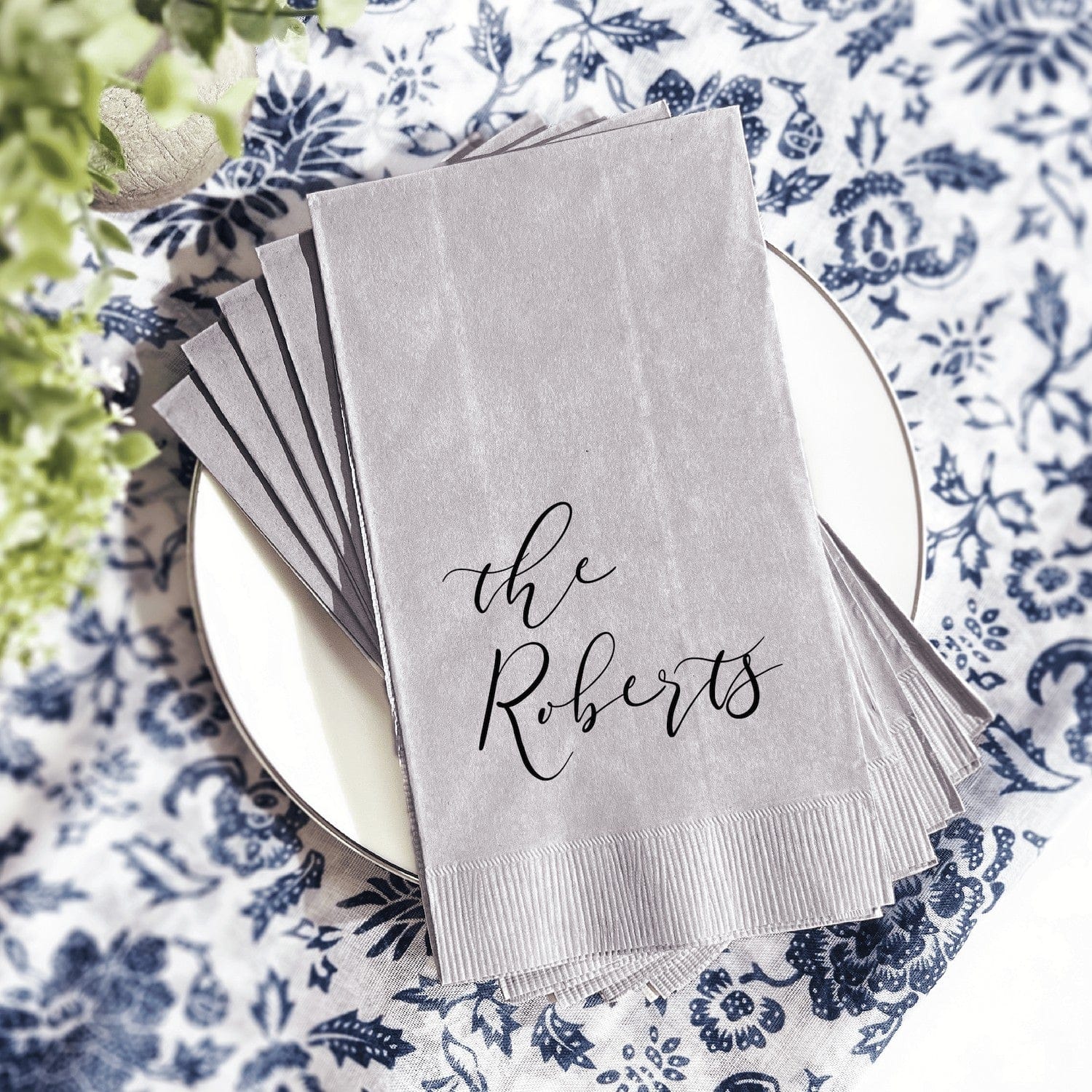 A stack of gray paper napkins with custom black script text, presented on a white plate atop a blue and white patterned tablecloth, perfect for personalized wedding or event table settings.