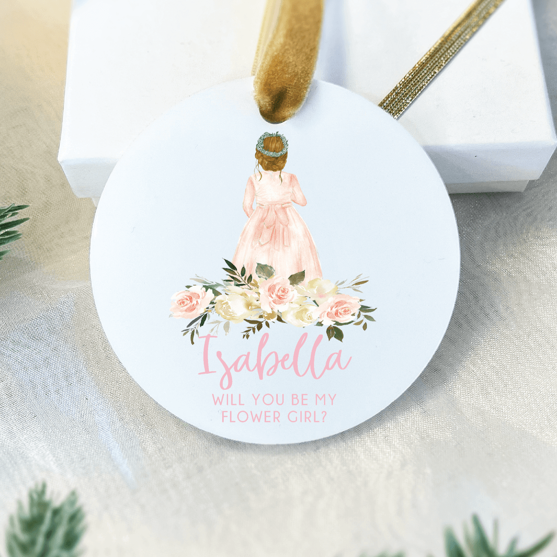 White round acrylic ornament with illustrated flower girl in pink dress, floral bouquet design, and personalized name "Isabella" with "Will you be my flower girl?" text below.