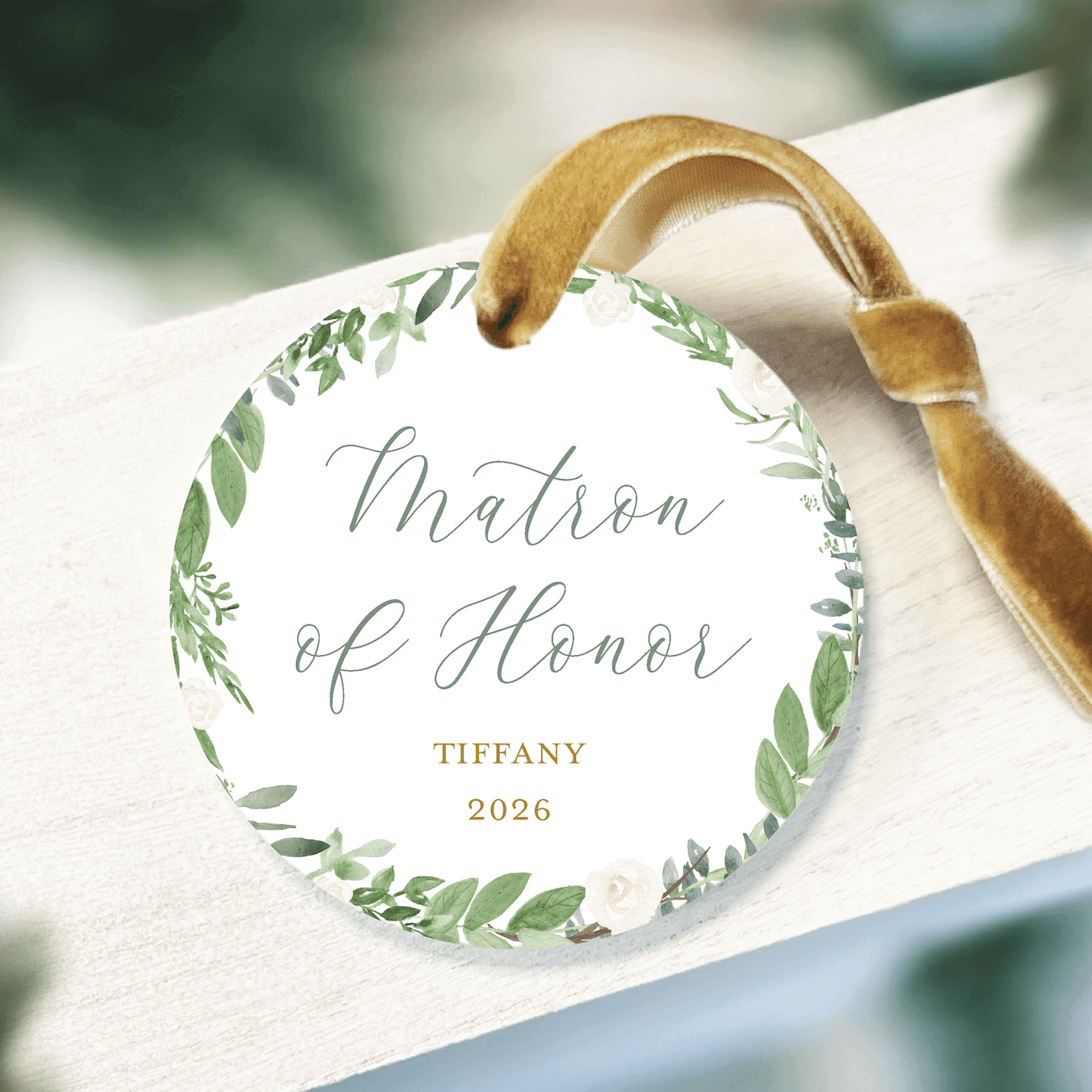 White acrylic ornament with green leaf wreath design, "Matron of Honor" text, personalized name "Tiffany" and year "2026" in gold, attached with tan ribbon.