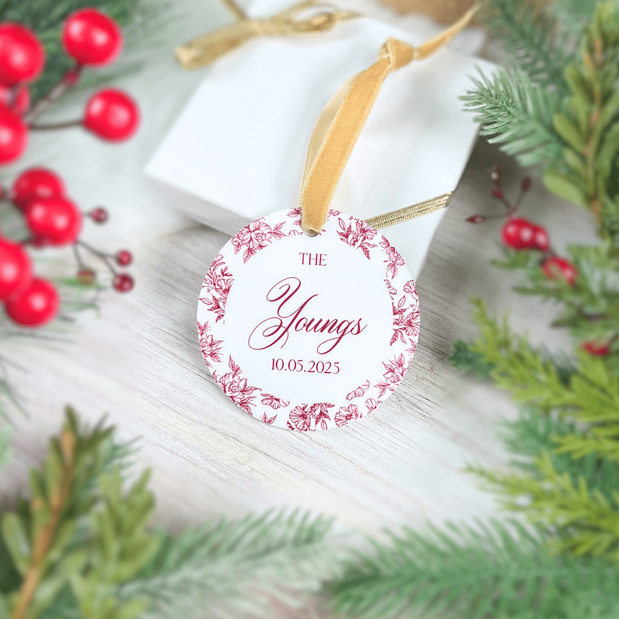 Round acrylic ornament with pink toile floral border, "The Youngs" text and date 8.10.2023, gold ribbon, displayed among evergreen branches and red berries.