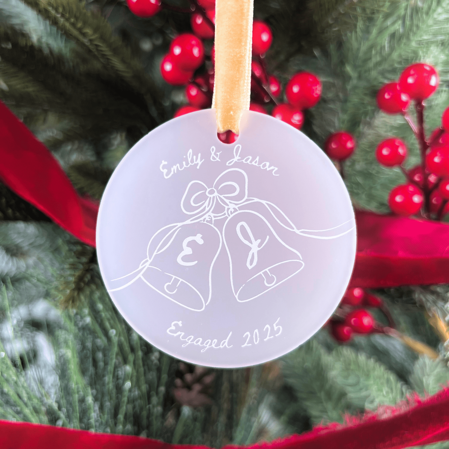 Frosted acrylic ornament with names "Emily & Jason" and wedding bells design, "Engaged 2025" engraved below, hanging from twine on festive greenery.