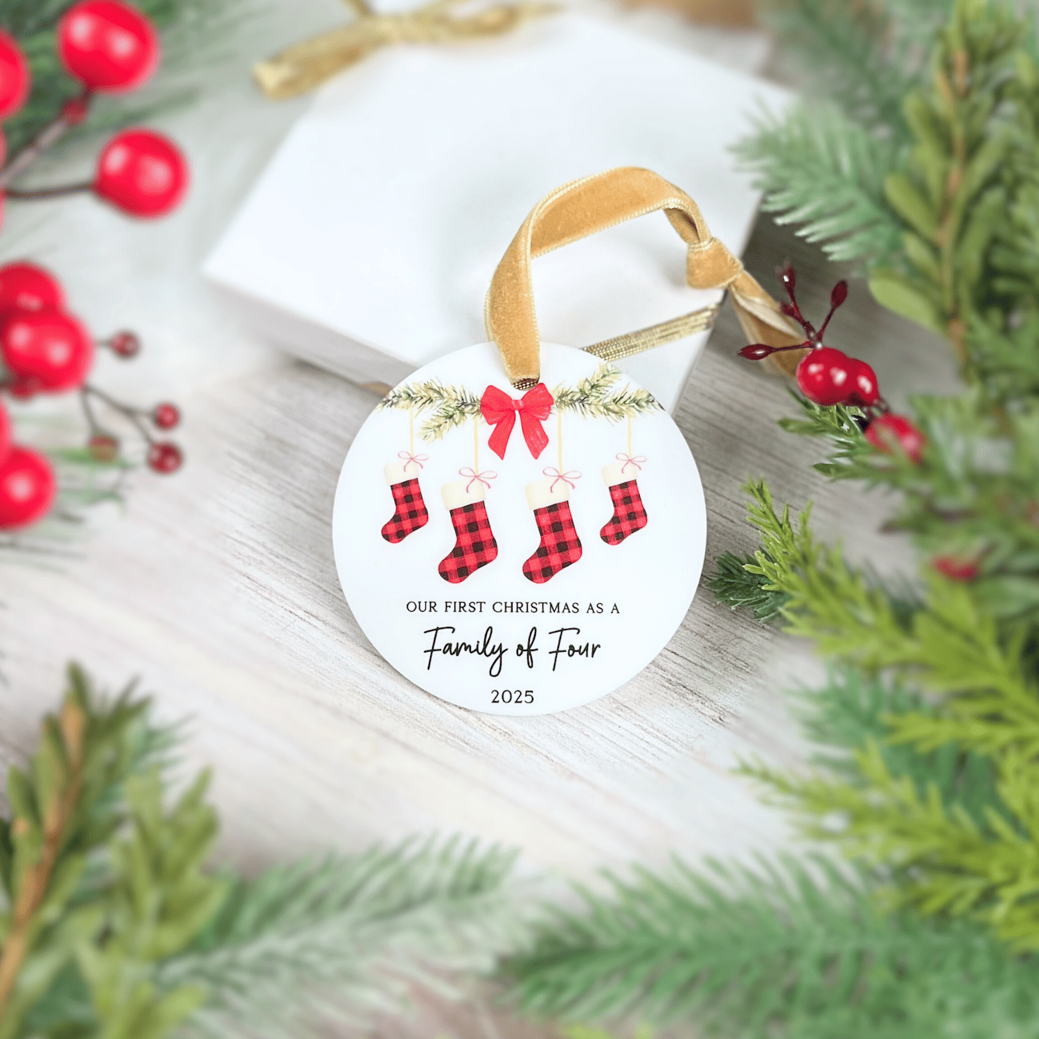 White acrylic ornament with gold ribbon, featuring four red stockings, holly garland, pink bow, and text reading "Our First Christmas as a Family of Four 2025"