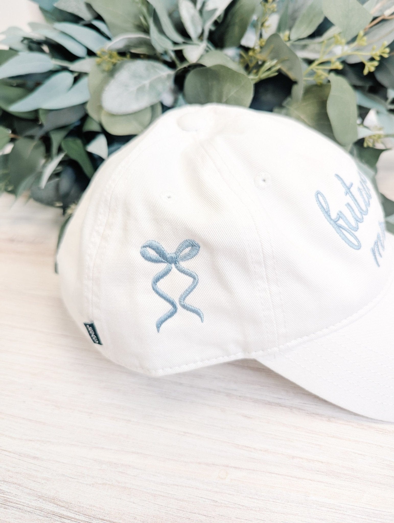 A white cotton baseball cap featuring light blue embroidered bow designs on the side, suitable for a wedding celebration or bridal shower.