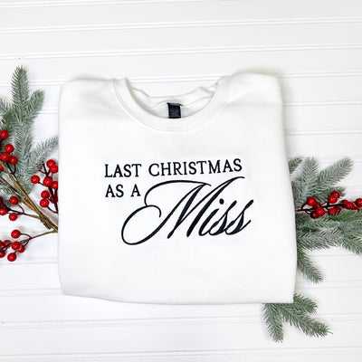 A white crewneck sweatshirt with black embroidered lettering is displayed on a white paneled surface, flanked by faux frosted pine sprigs and red berry accents.