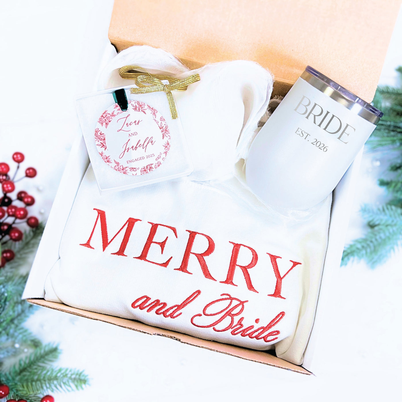 A festive white bridal gift box contains an insulated white tumbler with custom text, a cozy white embroidered sweatshirt, and a circular custom gift tag with a floral wreath design, ideal for a winter wedding or engagement celebration.