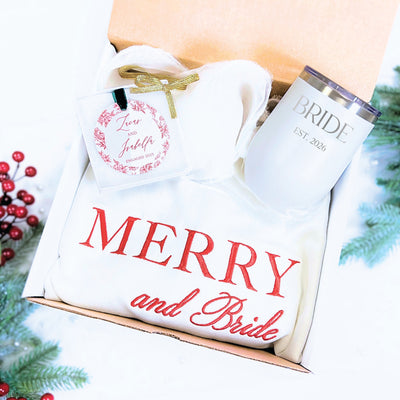 A festive white bridal gift box contains an insulated white tumbler with custom text, a cozy white embroidered sweatshirt, and a circular custom gift tag with a floral wreath design, ideal for a winter wedding or engagement celebration.