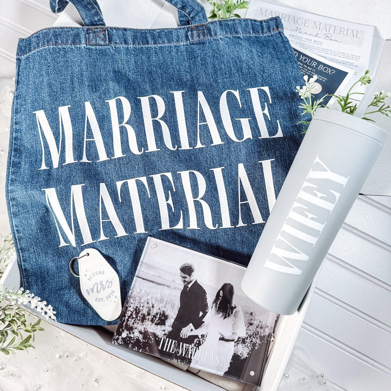 A curated bridal gift set featuring a blue denim tote bag, a light grey insulated tumbler, a white motel-style keychain, and a clear acrylic photo block, arranged with decorative greenery.