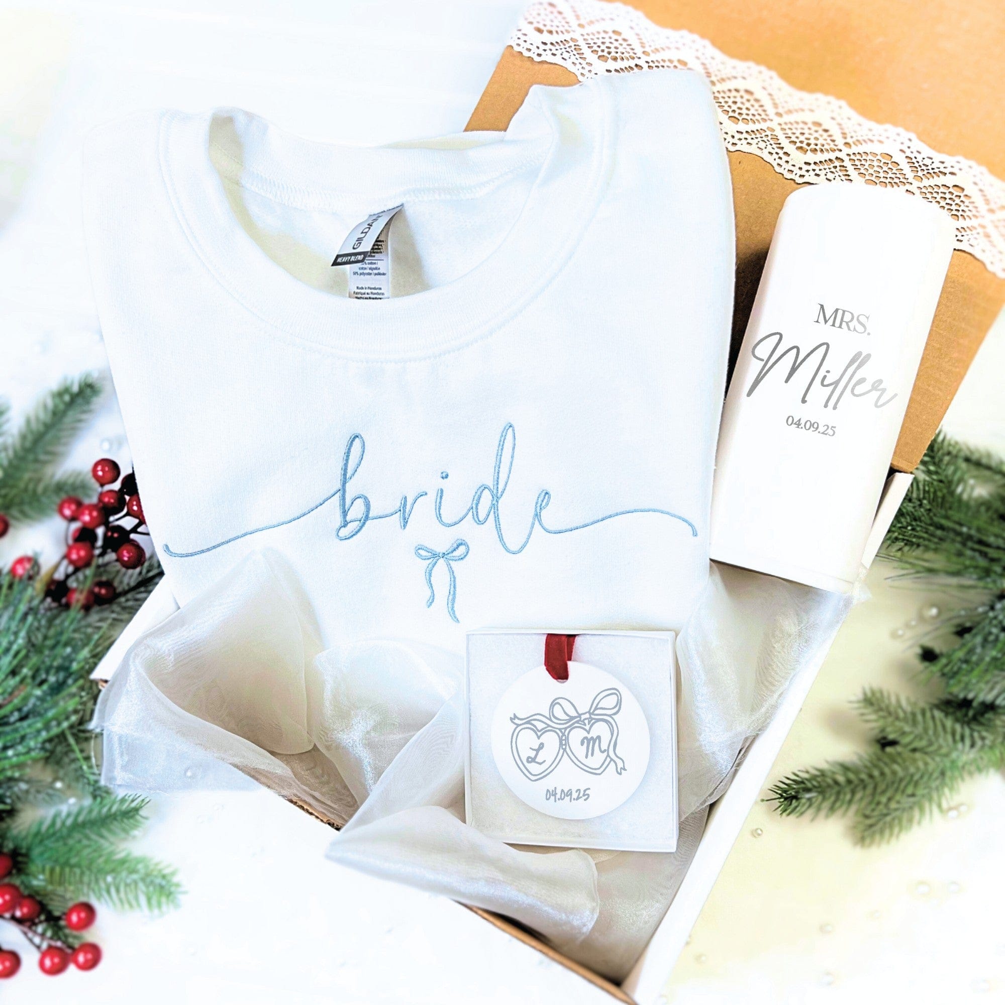 A white bridal gift set includes an embroidered sweatshirt, a personalized tumbler, and a personalized round ornament, all presented in a box with sheer fabric.
