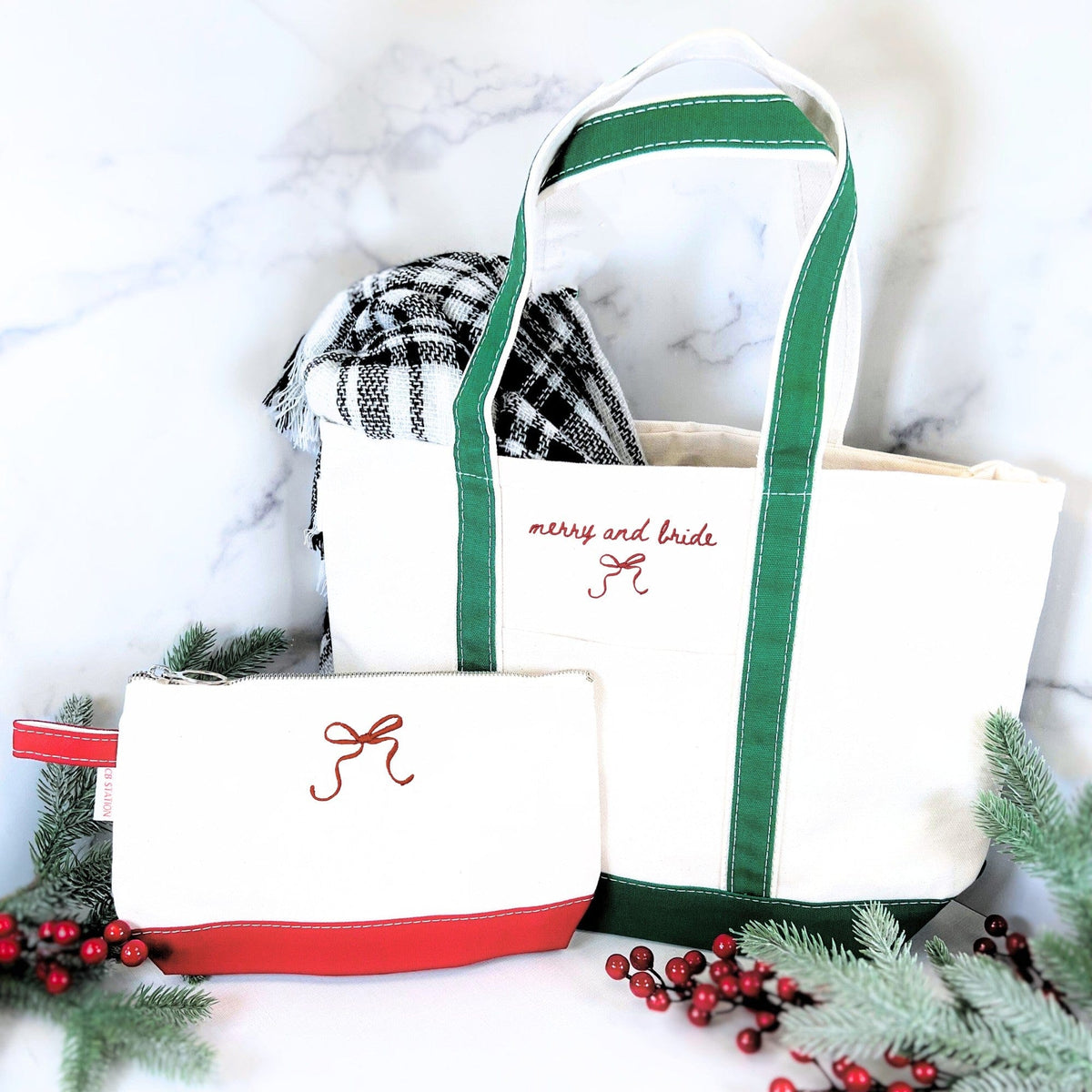 A white canvas tote bag with green straps and base, and a coordinating white canvas pouch with a red base, both embroidered with a decorative bow, are presented as a themed bridal gift set alongside a plaid blanket and holiday foliage.