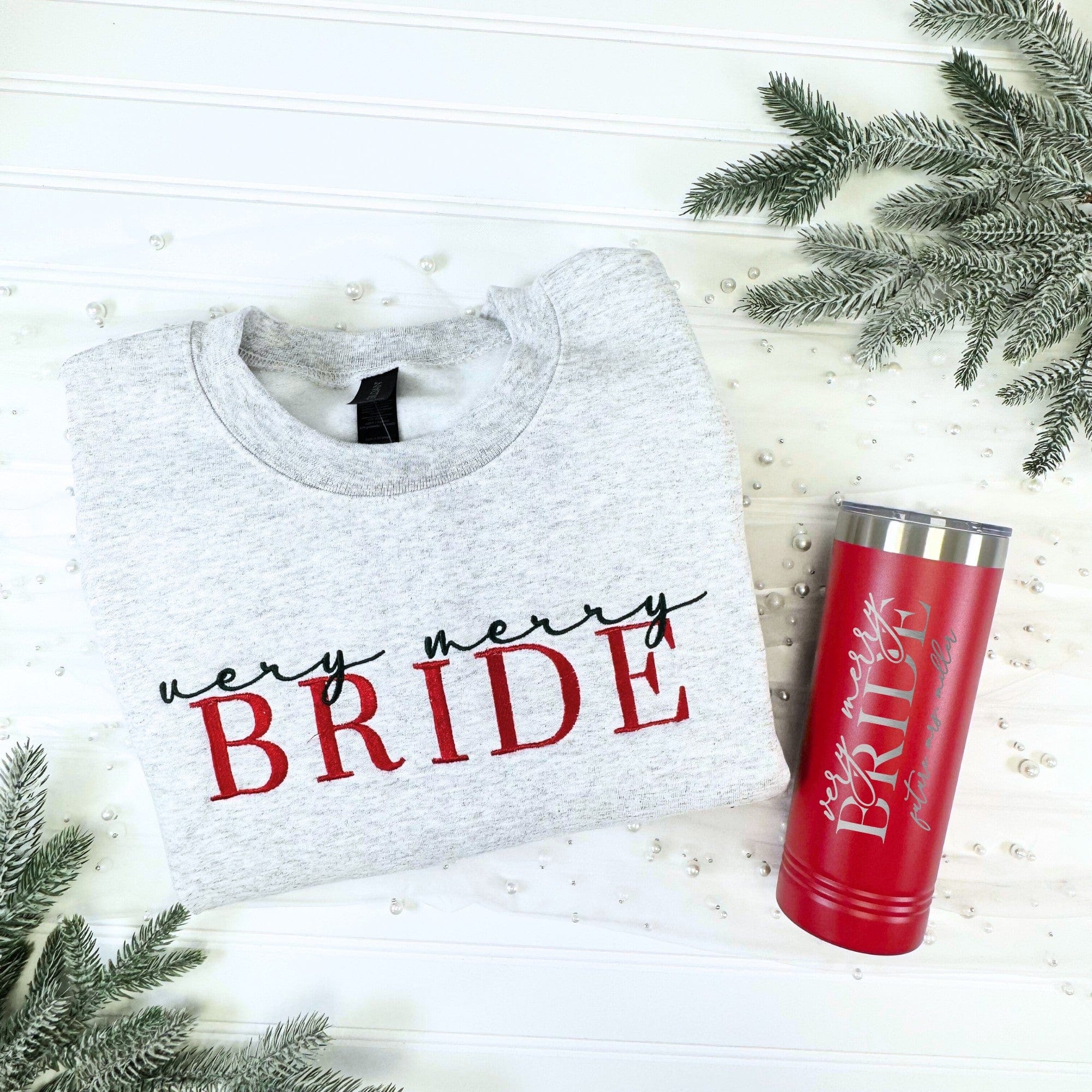 A grey cotton-blend sweatshirt with red and black embroidered text is paired with a red stainless steel insulated tumbler with silver engraved text, making a festive gift set for a winter wedding or bridal event.