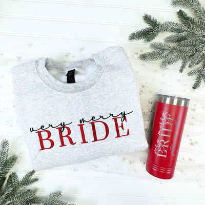 A grey cotton-blend sweatshirt with red and black embroidered text is paired with a red stainless steel insulated tumbler with silver engraved text, making a festive gift set for a winter wedding or bridal event.