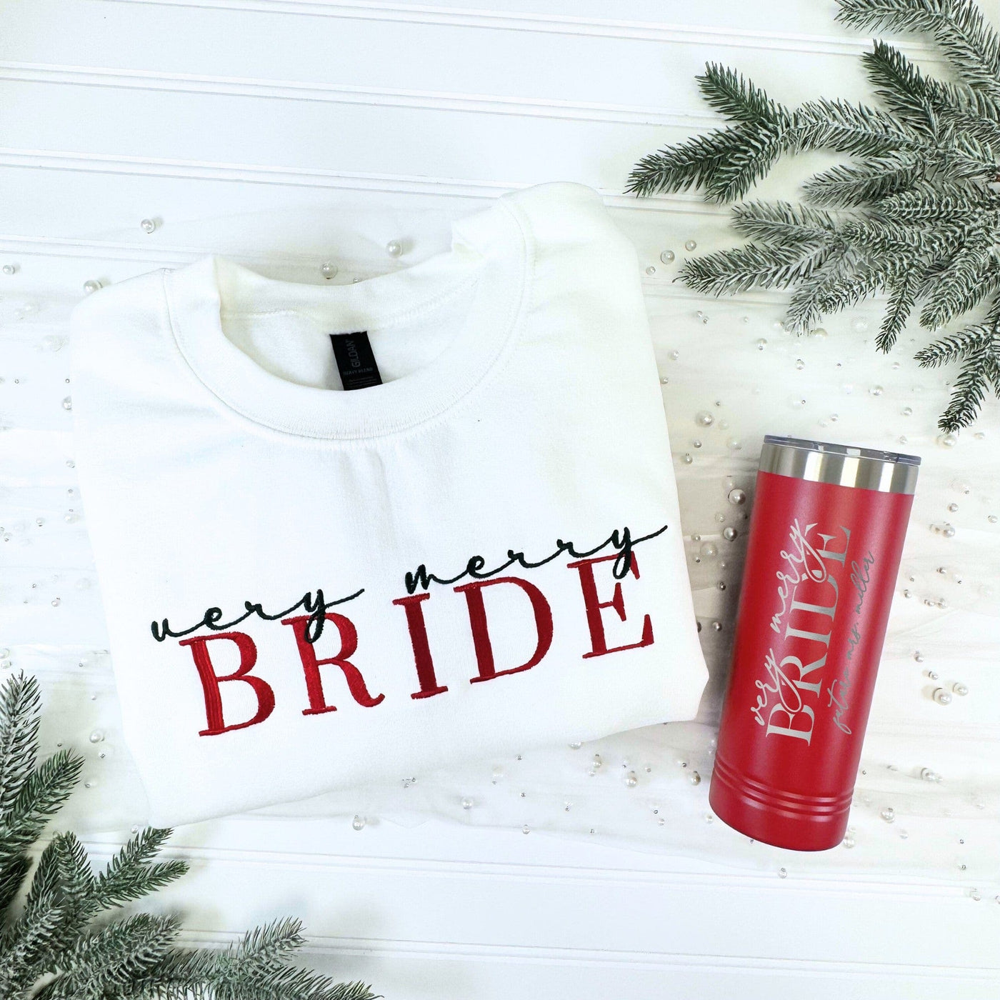 A white crewneck sweatshirt with festive red and black embroidery is displayed alongside a red stainless steel tumbler with coordinating silver engraving, presented for a winter bridal occasion.