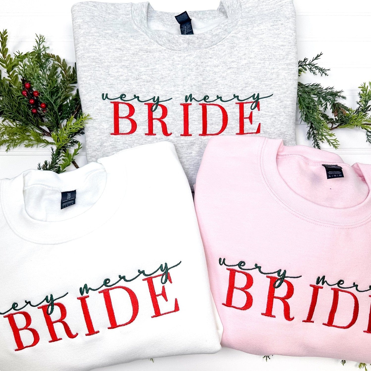 Three cozy crewneck sweatshirts in white, light gray, and pink feature embroidered green script and red block letters, ideal for a festive bridal occasion.
