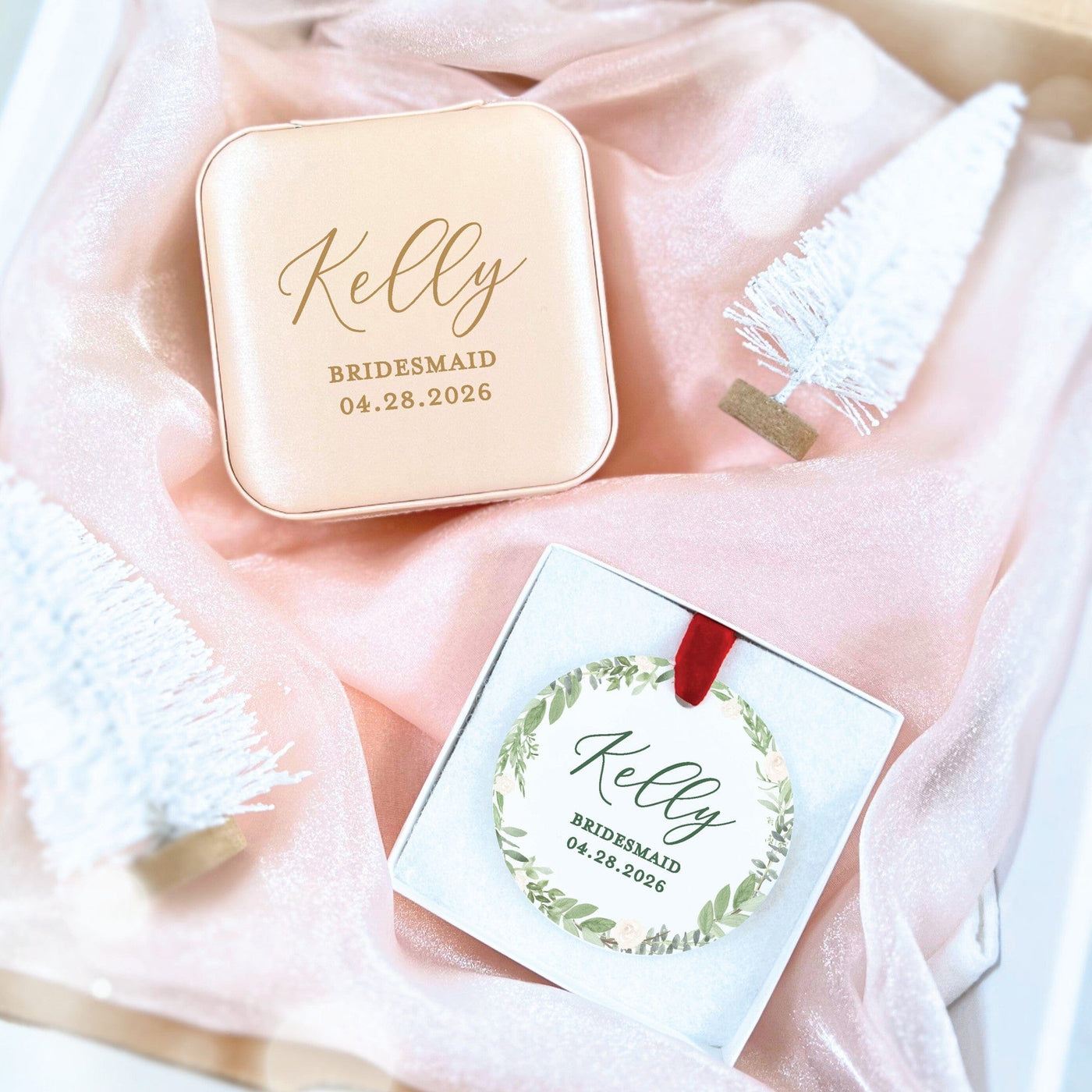 A personalized pale pink faux leather jewelry travel case and a round white ornament with a green floral wreath design and red ribbon, presented as a bridesmaid gift set on soft pink fabric.