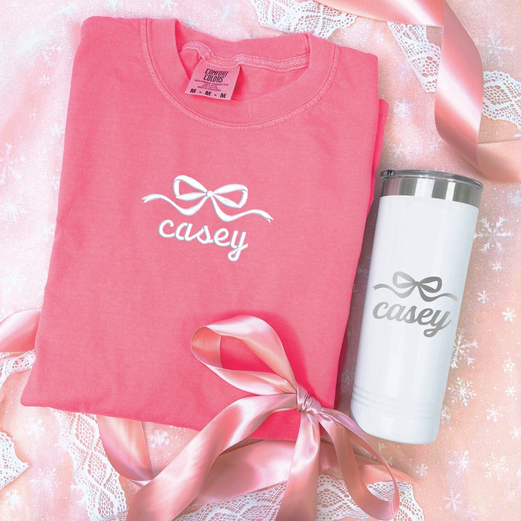 A coral pink short-sleeve shirt and a white insulated travel tumbler are displayed, both adorned with a decorative bow design, suitable as a personalized gift set for a bridal party.