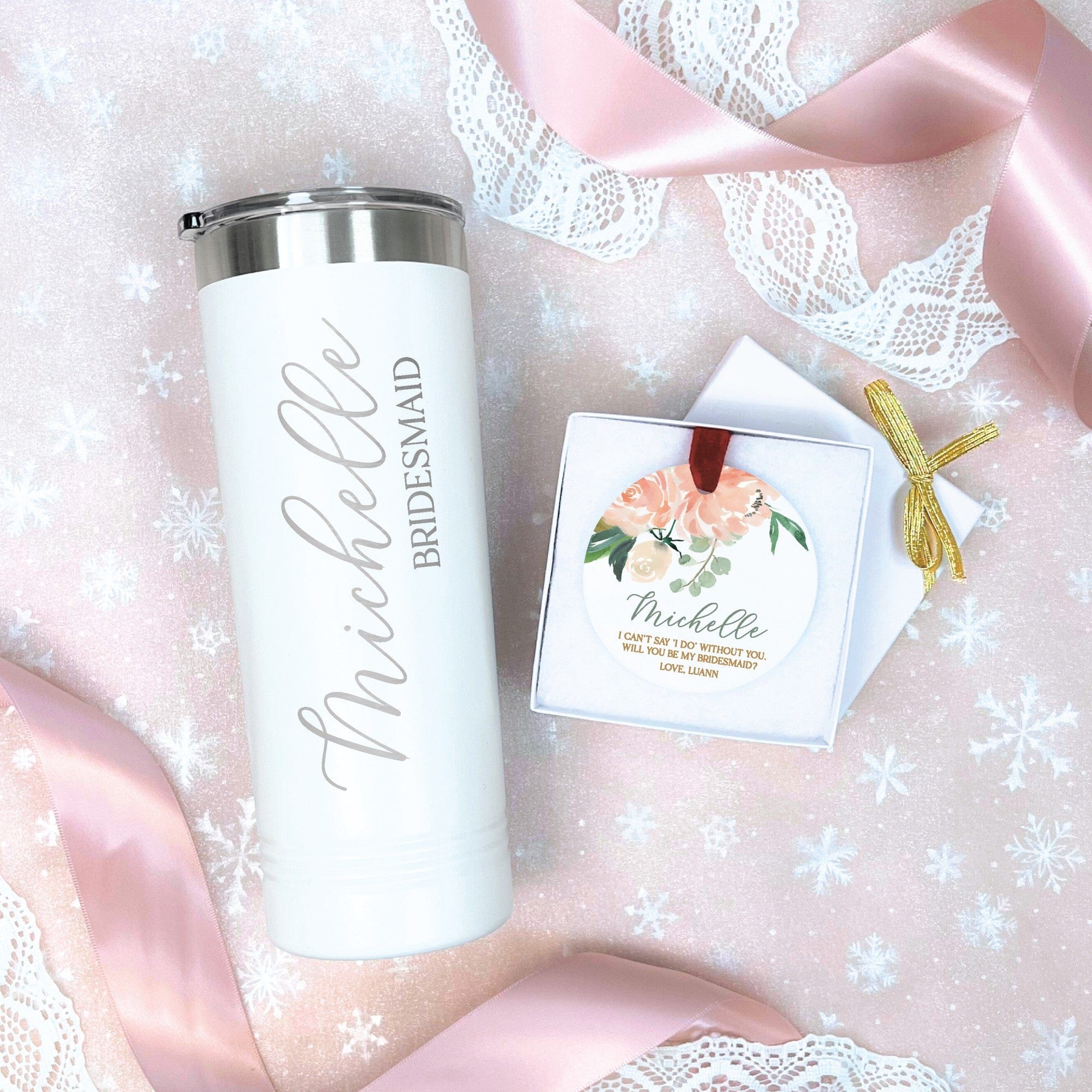 A personalized white insulated tumbler with a silver lid and a round ceramic ornament with a floral design are presented as a wedding party proposal gift set, arranged on a pink snowflake background with lace and satin ribbon.