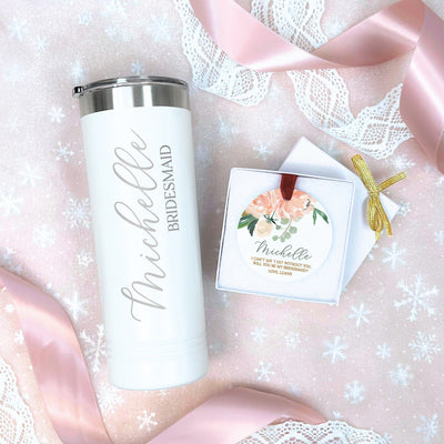 A personalized white insulated tumbler with a silver lid and a round ceramic ornament with a floral design are presented as a wedding party proposal gift set, arranged on a pink snowflake background with lace and satin ribbon.