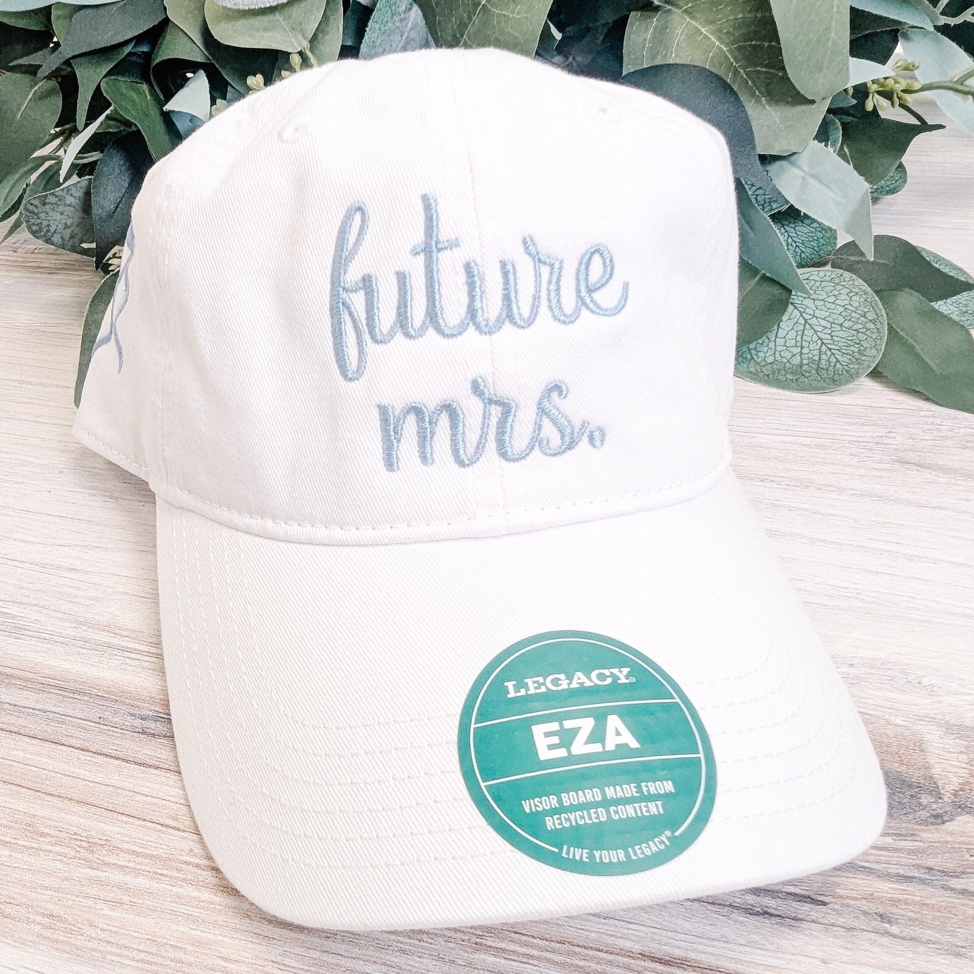 A white cotton baseball cap with light blue embroidered script and a tag noting the visor board is made from recycled content, designed for a bride-to-be.