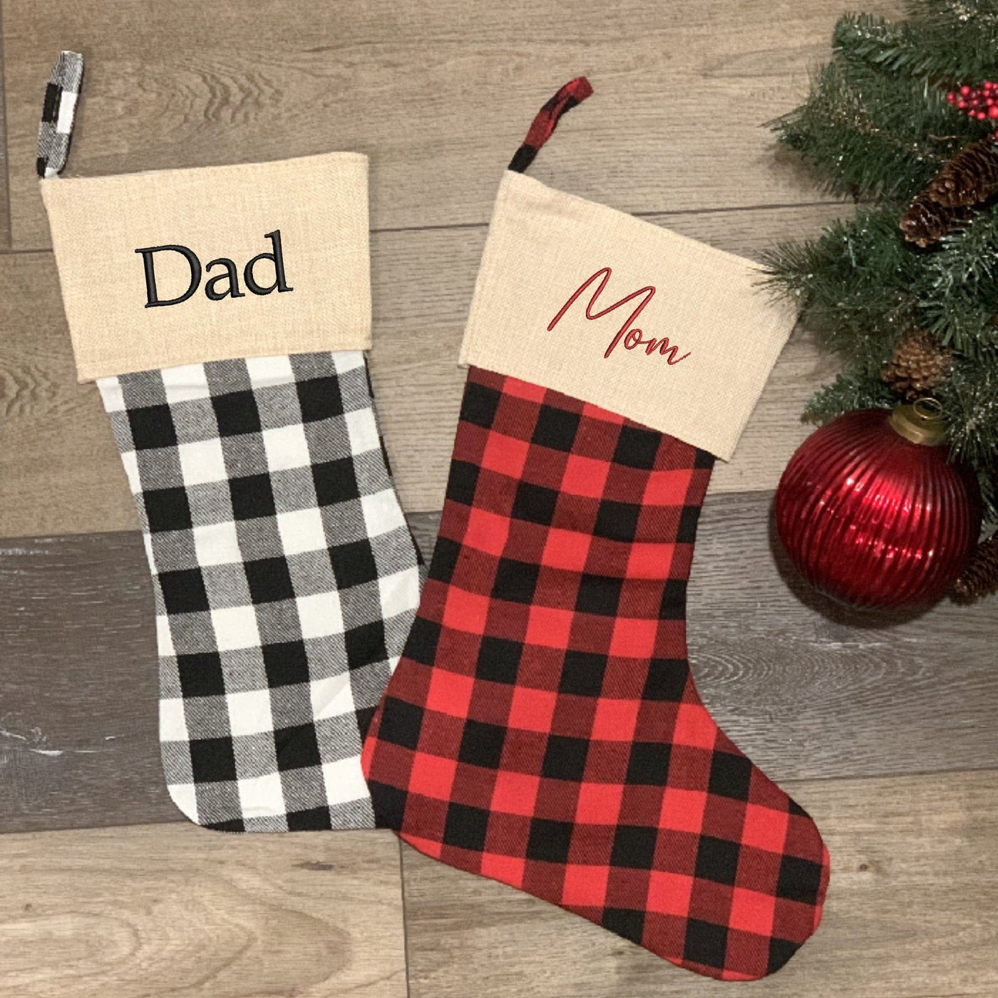 Two fabric Christmas stockings are displayed, one featuring black and white buffalo check plaid and the other red and black buffalo check plaid, both with a natural burlap-look cuff, designed for festive holiday decor and gift-giving.