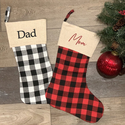 Two fabric Christmas stockings are displayed, one featuring black and white buffalo check plaid and the other red and black buffalo check plaid, both with a natural burlap-look cuff, designed for festive holiday decor and gift-giving.