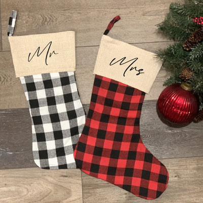 A pair of personalized Christmas stockings for holiday decor, one in black and white buffalo plaid and the other in red and black buffalo plaid, both featuring natural-colored fabric cuffs and hanging loops.