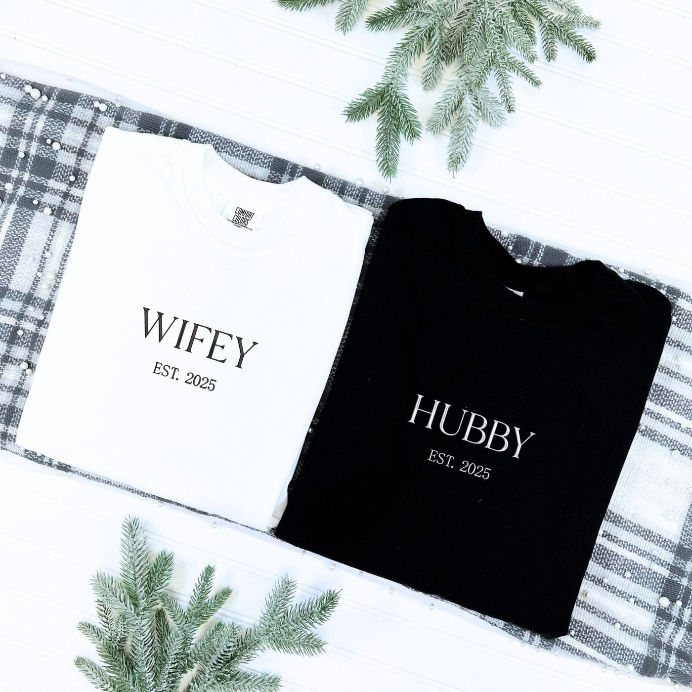 A personalized white cotton t-shirt and a black cotton t-shirt, custom designed for a couple, presented as a wedding or engagement gift set on a plaid fabric with pearl accents and frosted pine sprigs.
