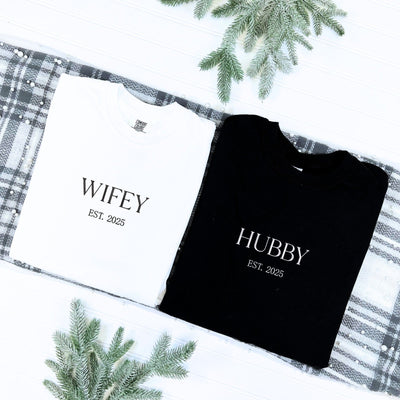A personalized white cotton t-shirt and a black cotton t-shirt, custom designed for a couple, presented as a wedding or engagement gift set on a plaid fabric with pearl accents and frosted pine sprigs.
