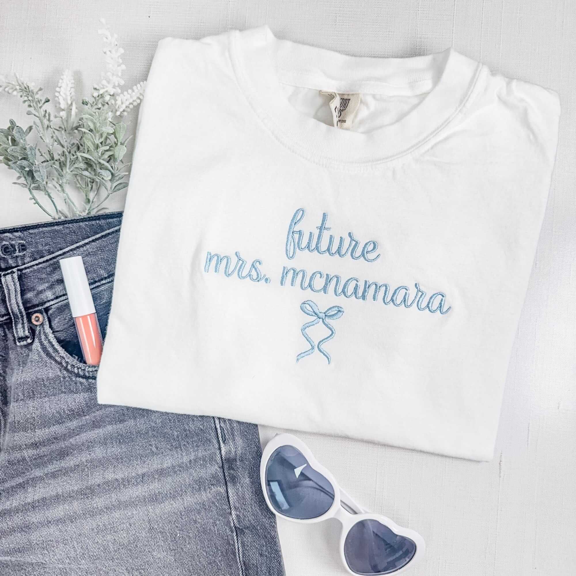 A soft white t-shirt featuring light blue script embroidery and a delicate bow design, perfect for pre-wedding celebrations.